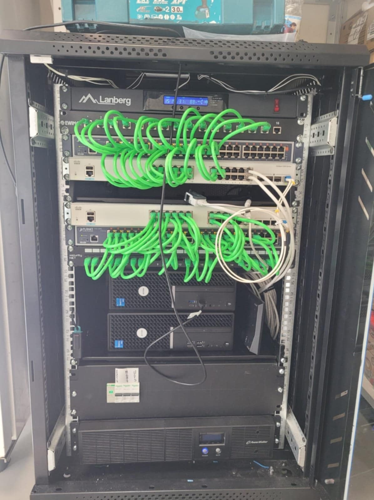 Szafa rackowa z zamontowanymi urządzeniami sieciowymi: switche, routery, serwery i zasilacz awaryjny, połączone zielonymi kablami patchcord, widoczne oznaczenia Lanberg i Planet.