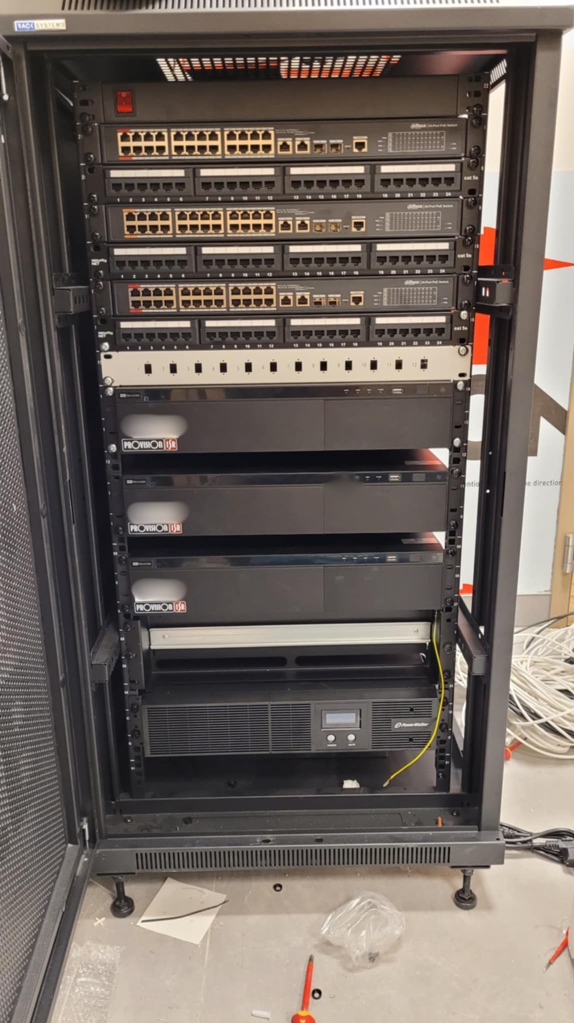 Szafa rack z zainstalowanymi przełącznikami sieciowymi, rejestratorami Provision ISR i zasilaczem awaryjnym PowerWalker, widoczne kable i narzędzia na podłodze.
