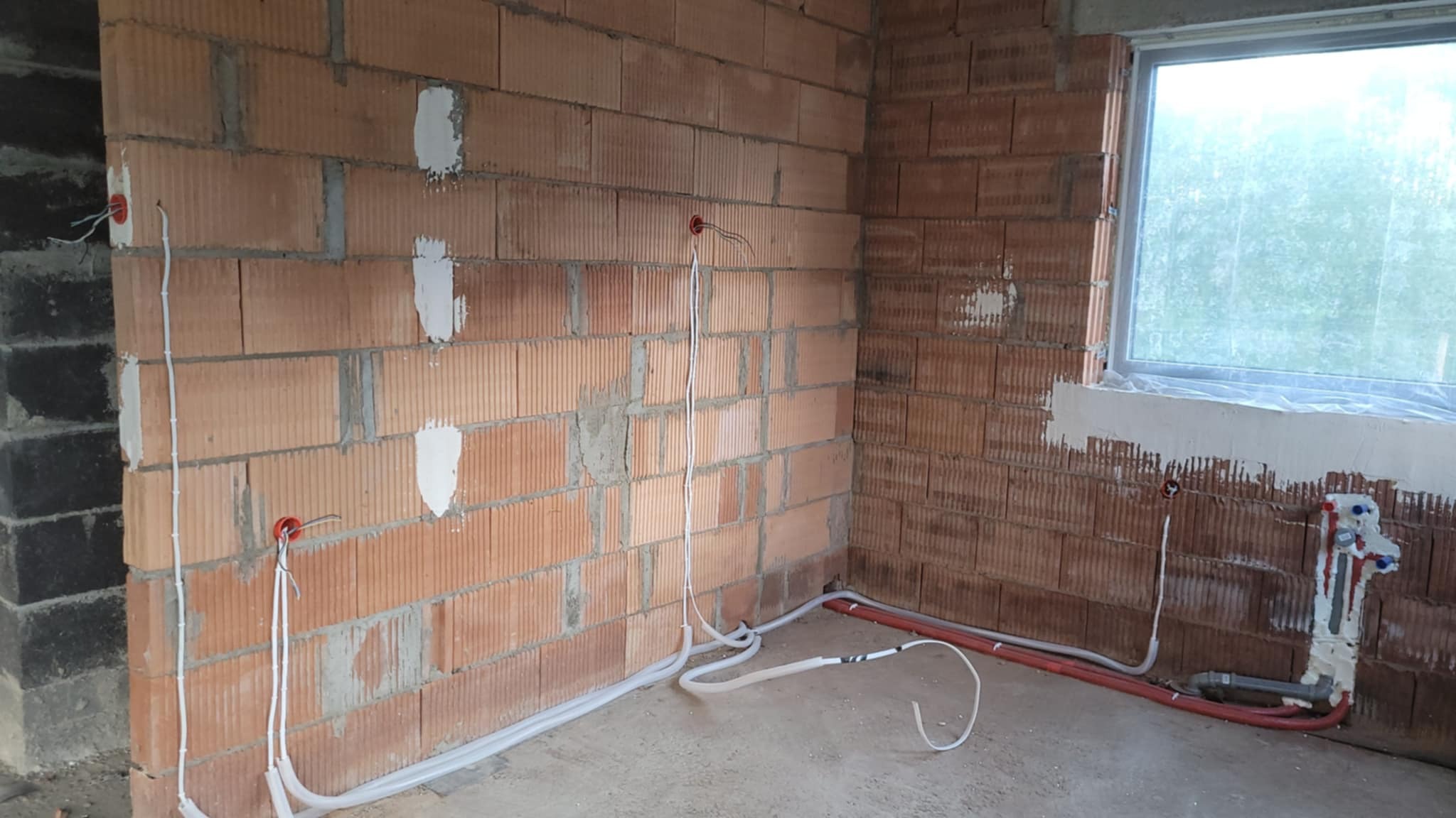 Surowy mur z czerwonej cegły z widocznymi przewodami elektrycznymi w białych rurkach i puszkami instalacyjnymi w kolorze pomarańczowym; widoczne także rury wodne i okno z folią ochronną.