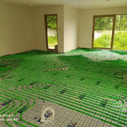 Proinstal - Zielone rury systemu rozprowadzenia ciepła podłogowego ułożone na siatce w jasnym, pustym pokoju z dużymi oknami wychodzącymi na zieleń.