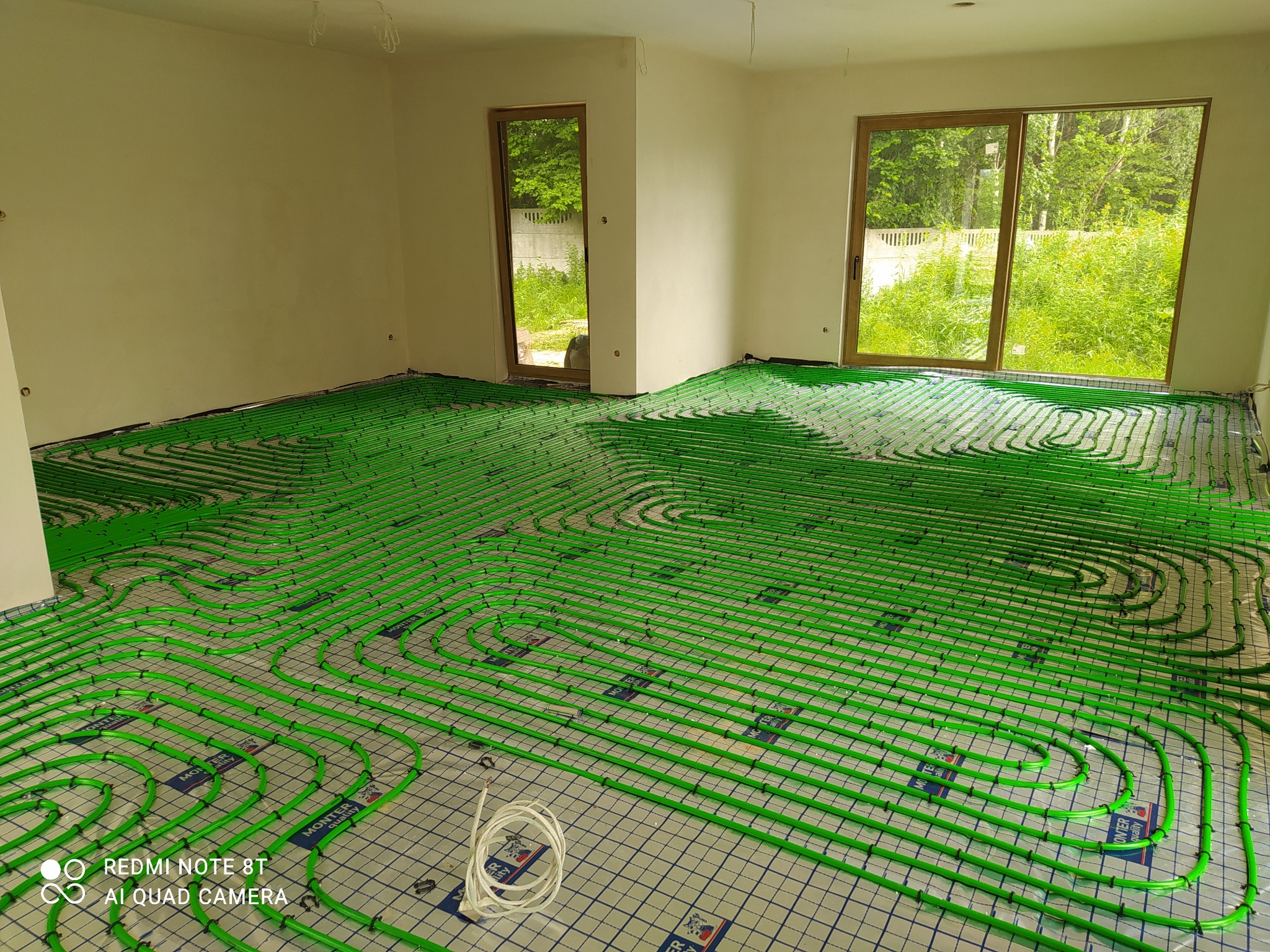 Zielone rurki ogrzewania podłogowego ułożone na podkładzie izolacyjnym w jasnym, nowym pomieszczeniu z dużymi oknami wychodzącymi na zieleń.