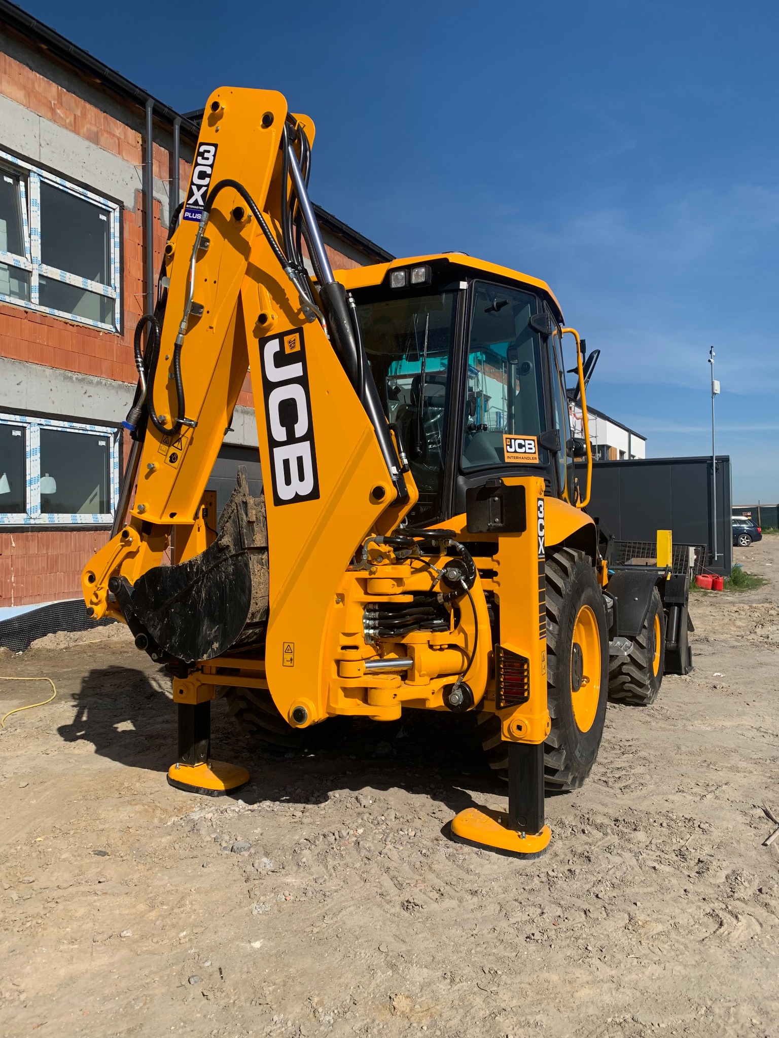 Żółta koparko-ładowarka JCB 3CX zaparkowana na placu budowy, z widocznym nowym budynkiem w trakcie wznoszenia w tle, stabilizowana na wysuwanych podporach.