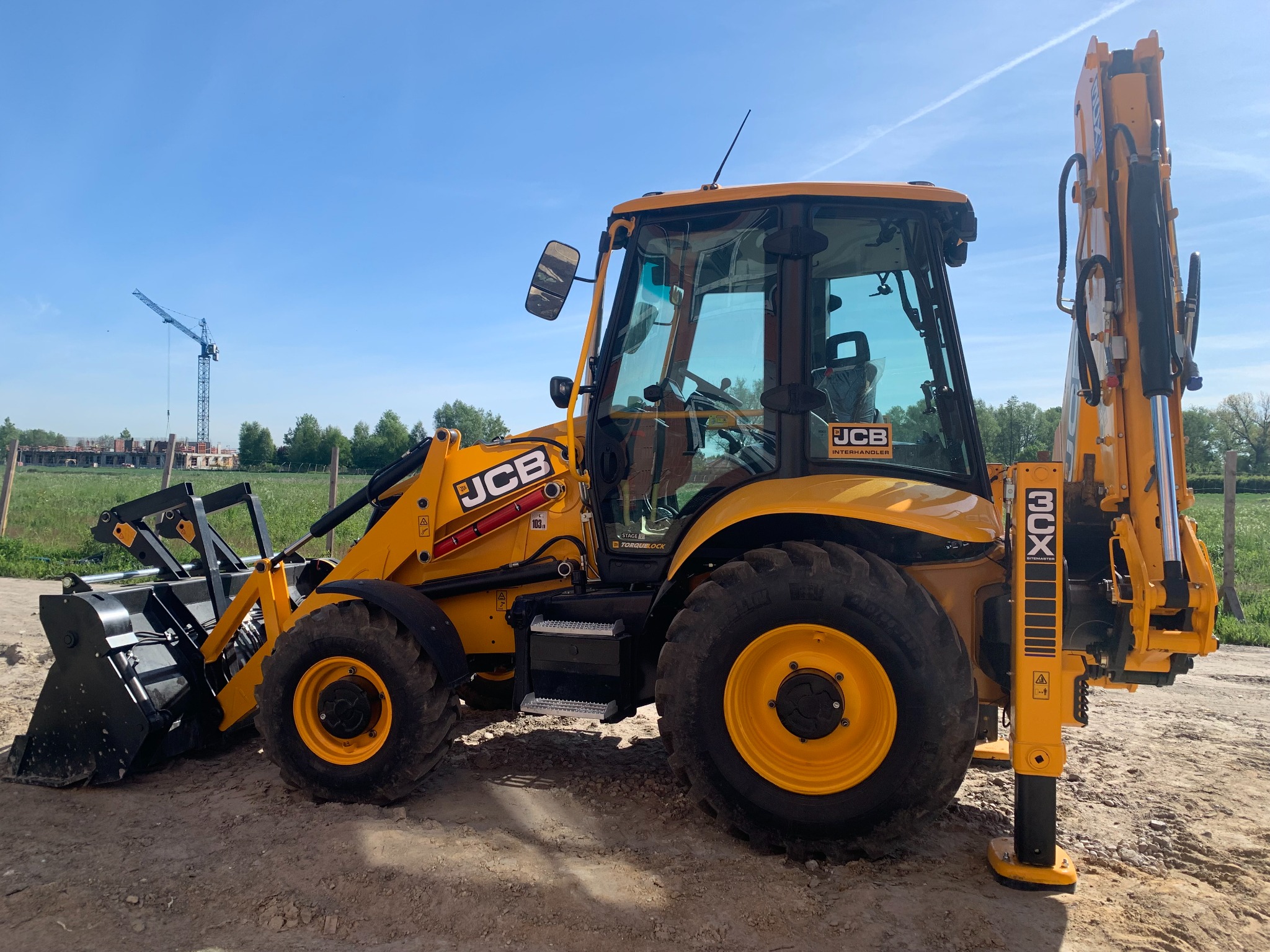 Żółta koparko-ładowarka JCB 3CX zaparkowana na placu budowy, z widocznym w tle żurawiem budowlanym i zielenią.