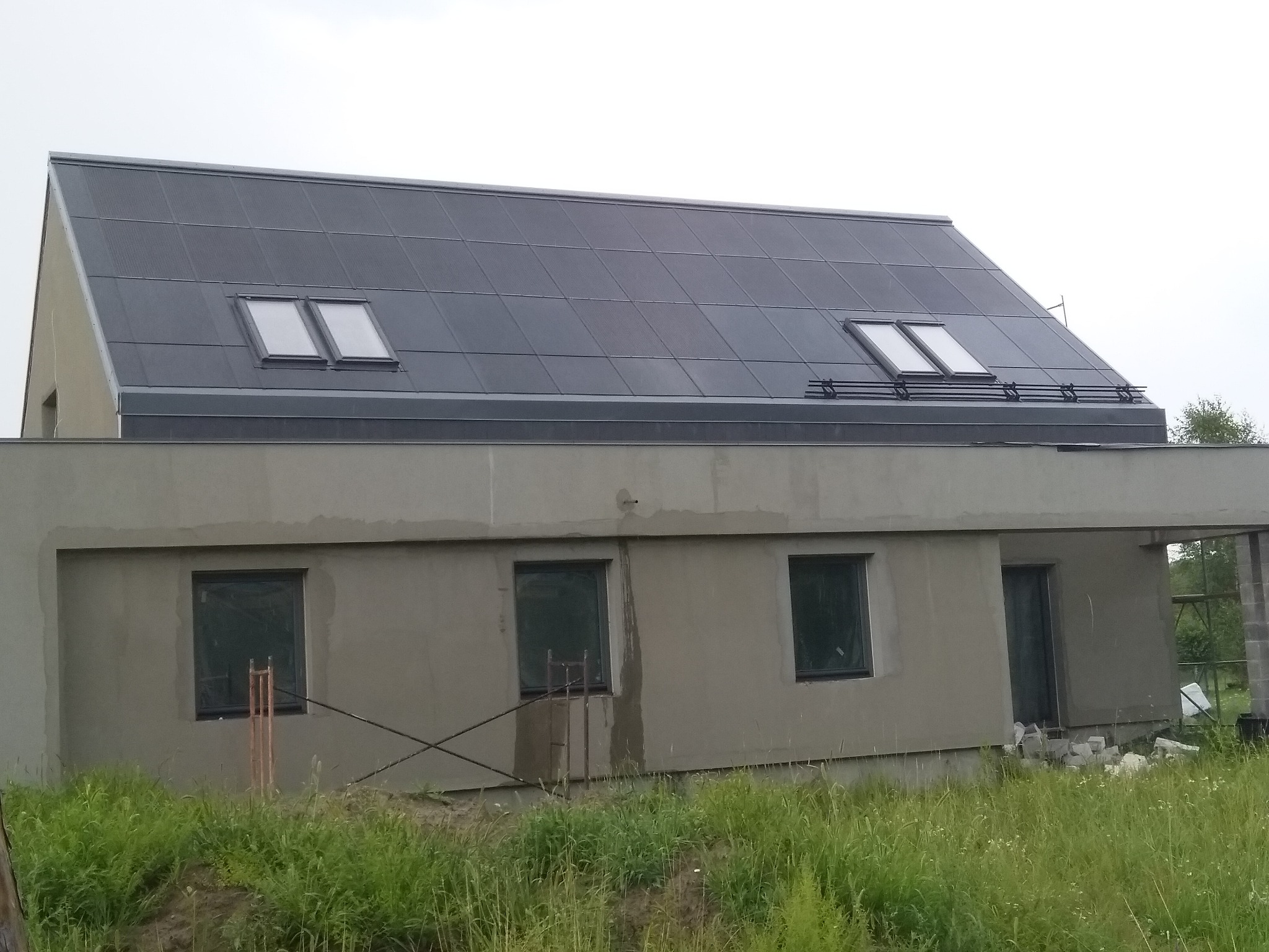 Dom w trakcie budowy z zamontowanym ciemnoszarym dachem z blachy z dwoma oknami dachowymi, widoczne rusztowania i materiały budowlane wokół budynku.
