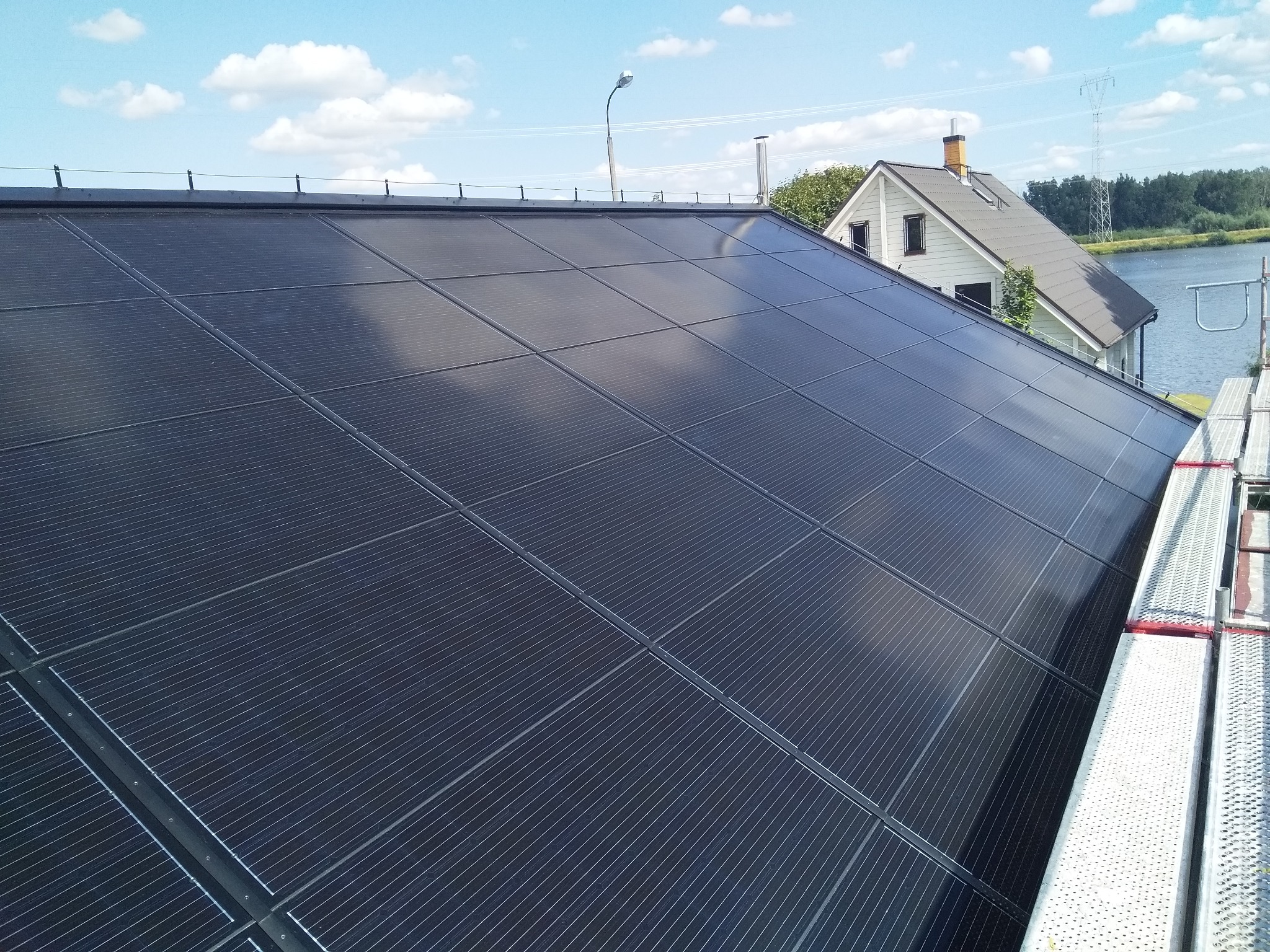 Nowoczesny, czarny dach z panelami słonecznymi na tle błękitnego nieba z chmurami, widoczny fragment rusztowania i dom z kominem w tle.