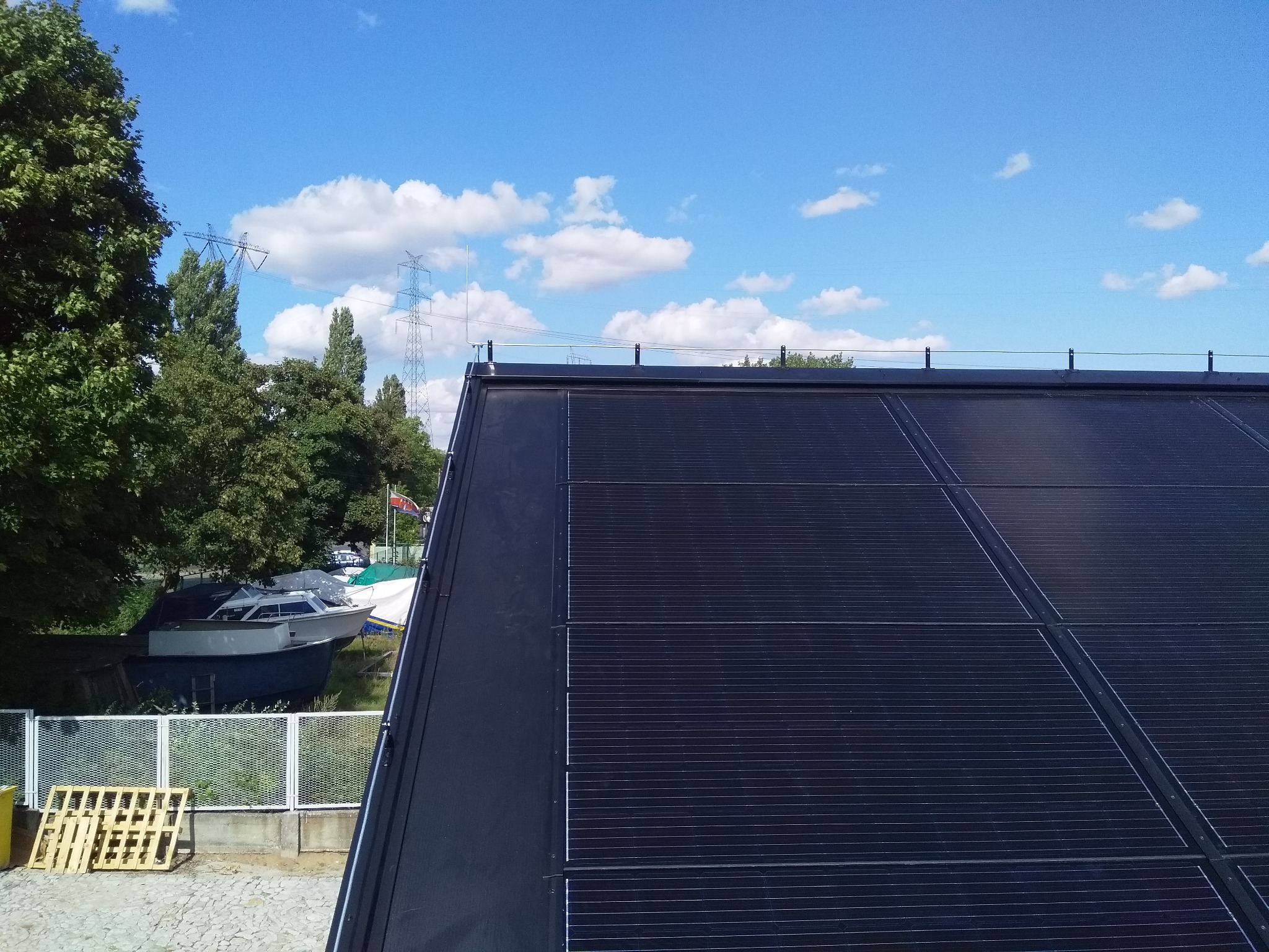 Czarny dach z panelami fotowoltaicznymi widziany z góry, w tle drzewa, łodzie i błękitne niebo z chmurami, w rogu widoczne palety.