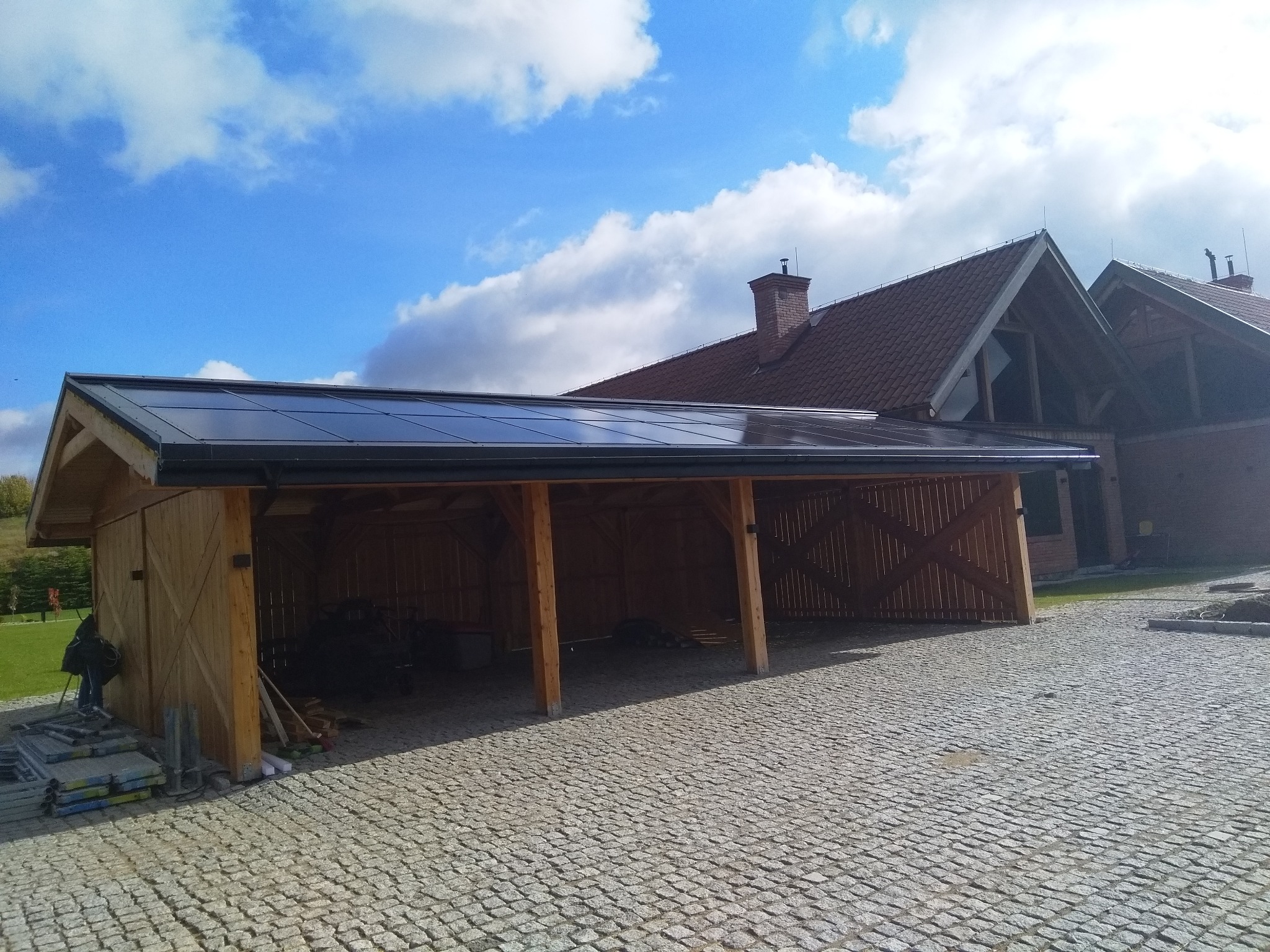 Drewniana wiata garażowa z panelami fotowoltaicznymi na dachu, widok z zewnątrz na brukowanym podjeździe, budynek mieszkalny w tle, częściowo pochmurne niebo.