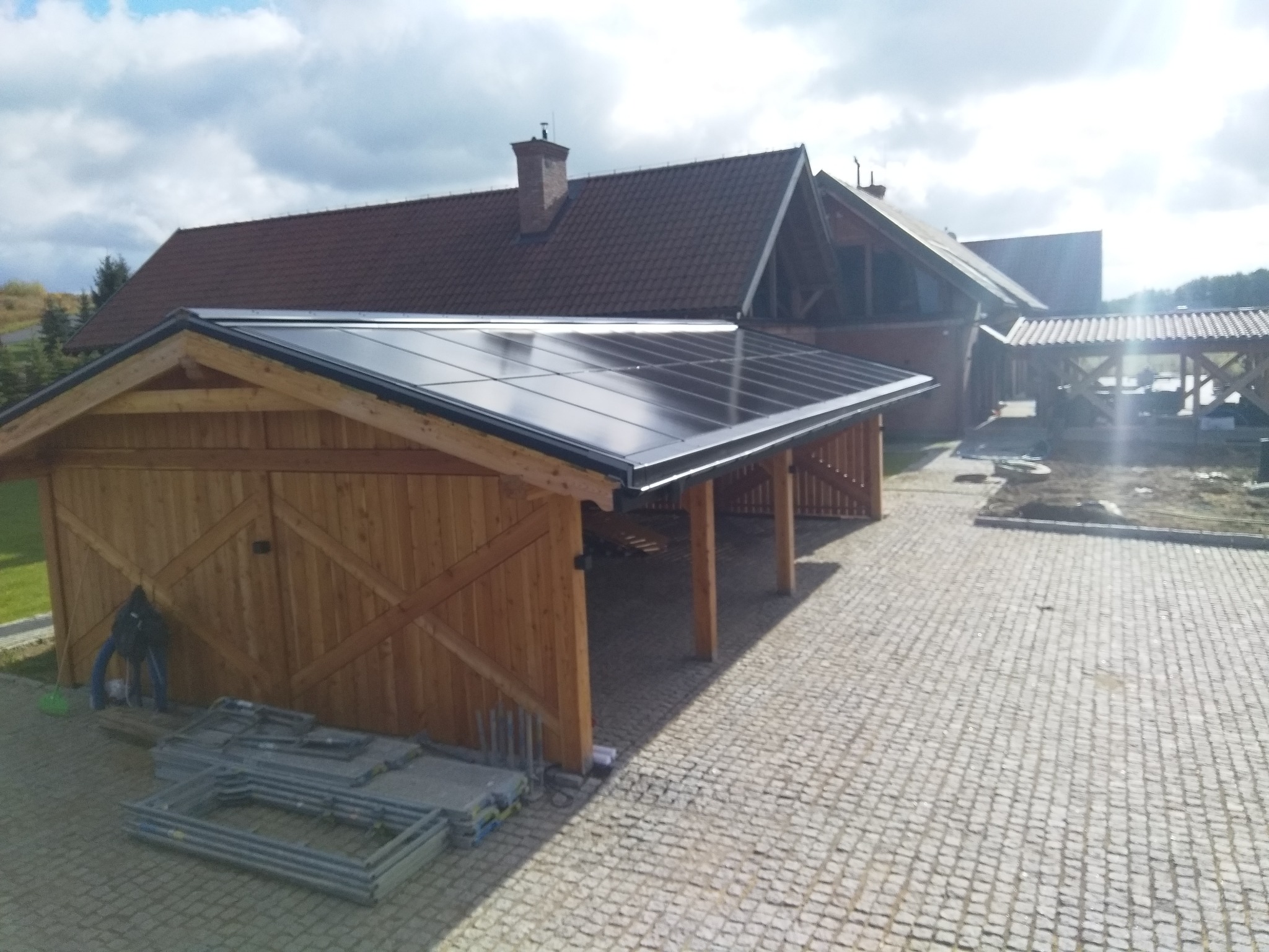Drewniana wiata garażowa z panelami słonecznymi na dachu, na pierwszym planie ułożone rusztowania, w tle budynek z brązowym dachem z dachówki.