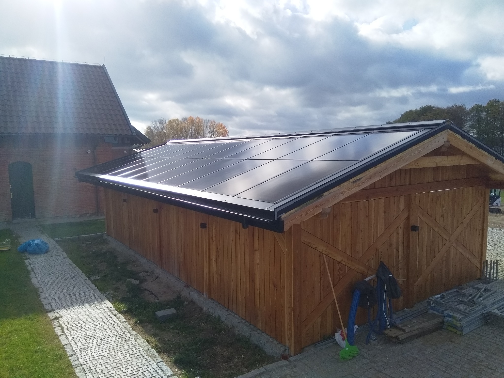 Nowoczesny budynek gospodarczy z drewnianą elewacją i panelami fotowoltaicznymi na dachu, widok z góry, obok kostka brukowa i budynek z czerwonej cegły.