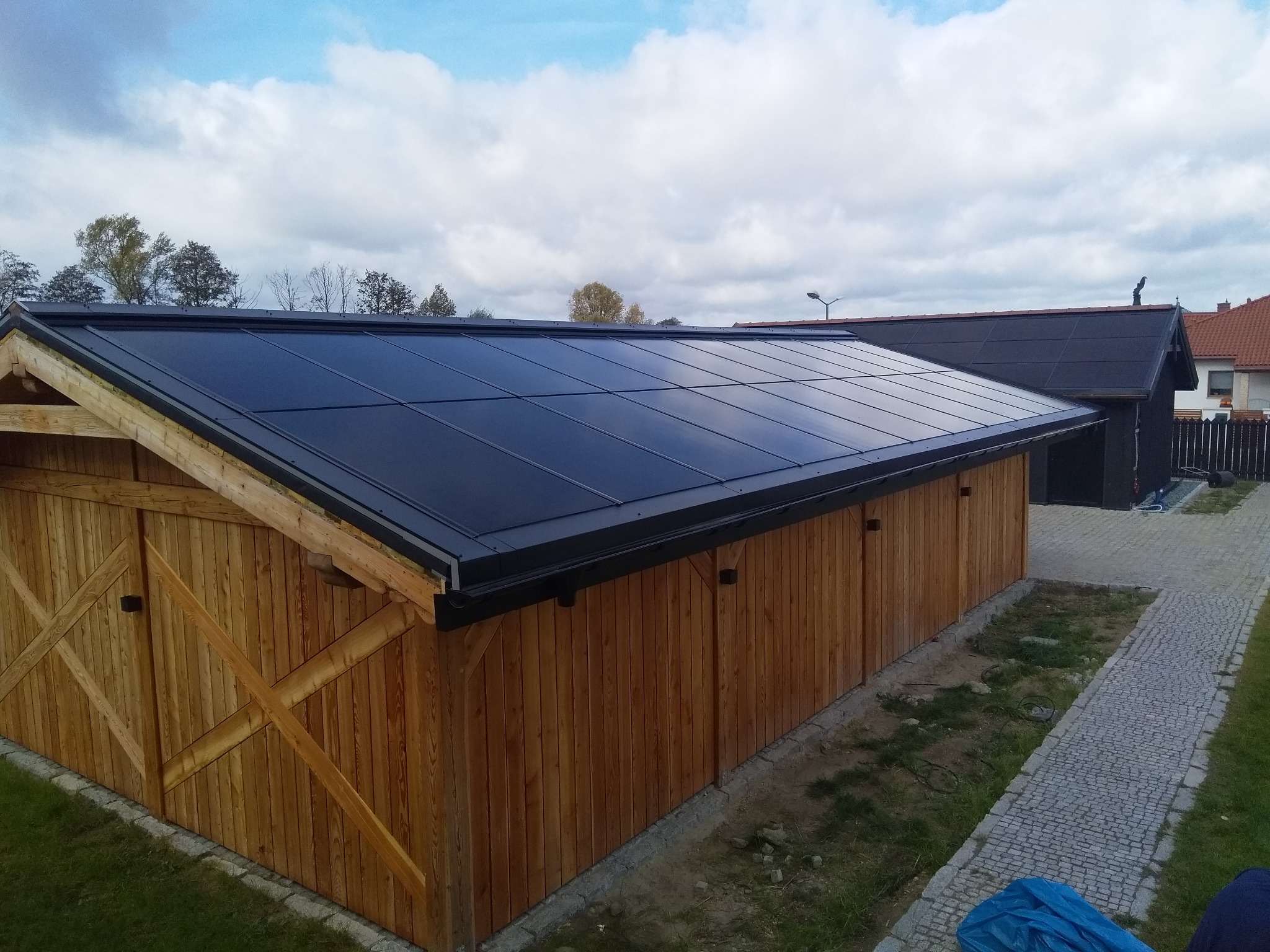 Nowoczesny, czarny dach z blachy trapezowej na drewnianym budynku gospodarczym z widocznym orynnowaniem i kostką brukową w tle.