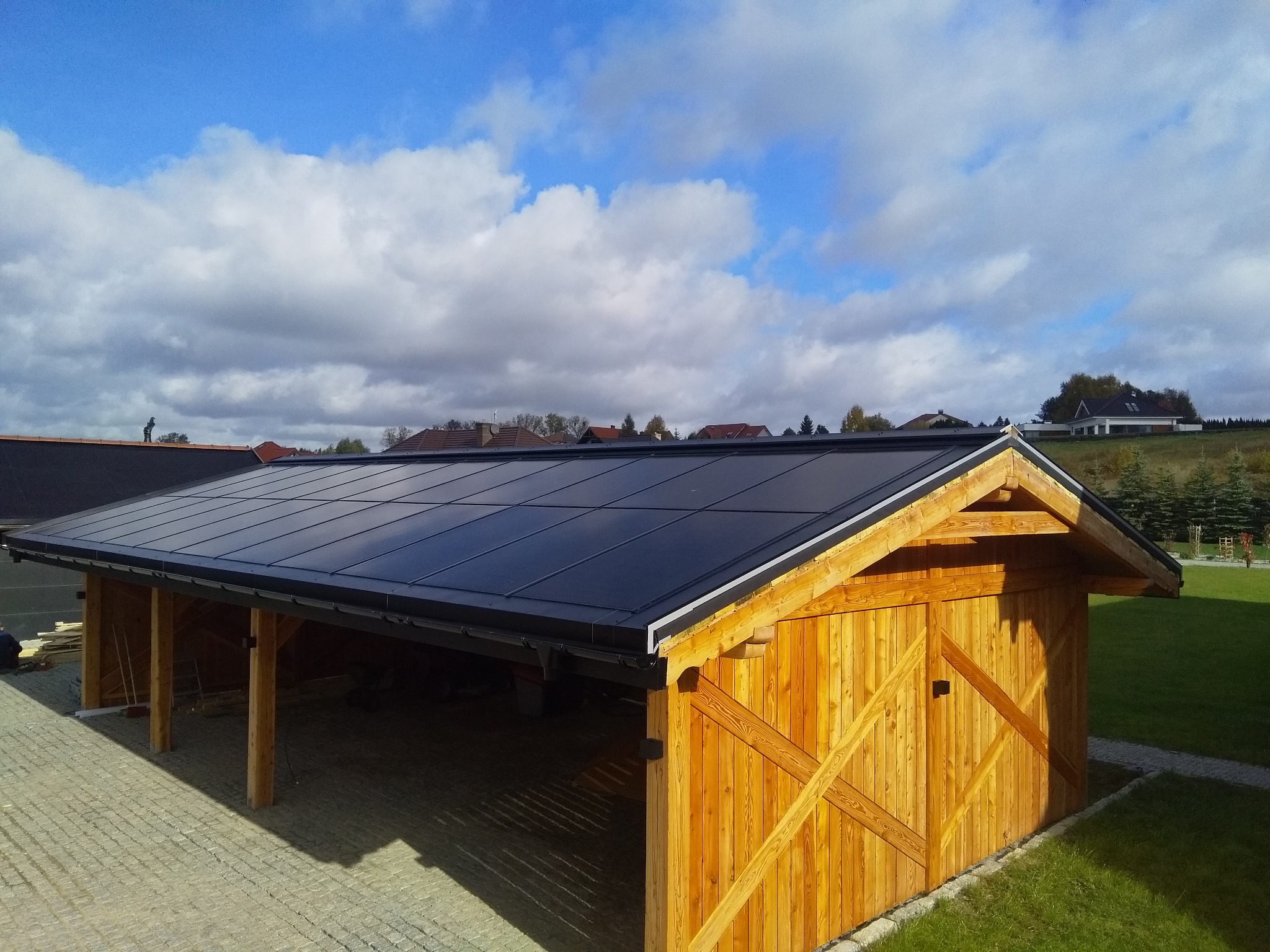 Nowoczesna wiata garażowa z drewnianą konstrukcją i panelami fotowoltaicznymi na dachu, na tle błękitnego nieba z chmurami.