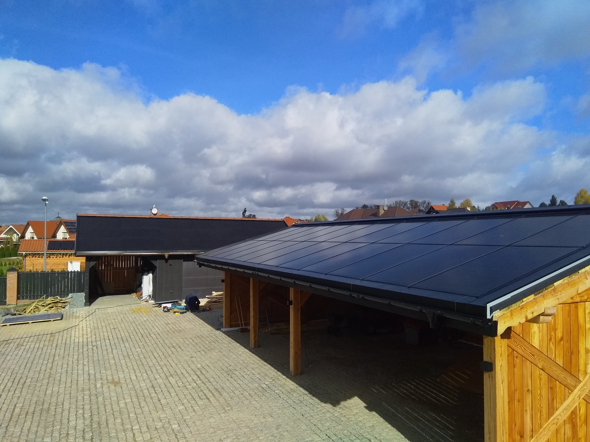 Widok z góry na drewnianą konstrukcję wiaty z czarnym, płaskim pokryciem dachowym; w tle domy jednorodzinne i błękitne niebo z chmurami.