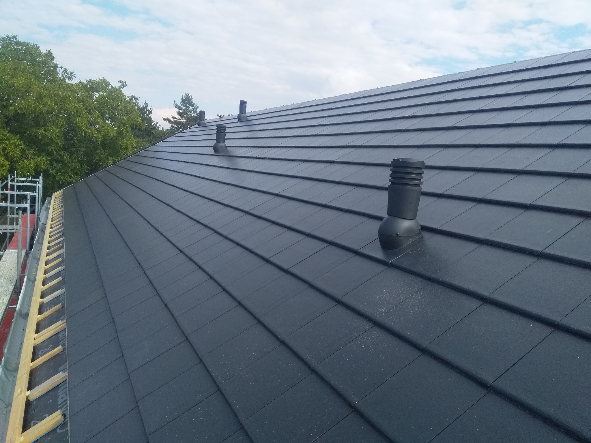 Nowy, ciemny dach z ceramicznych płytek z kominkami wentylacyjnymi, widok z góry, częściowo widoczne rusztowanie i drzewa w tle.