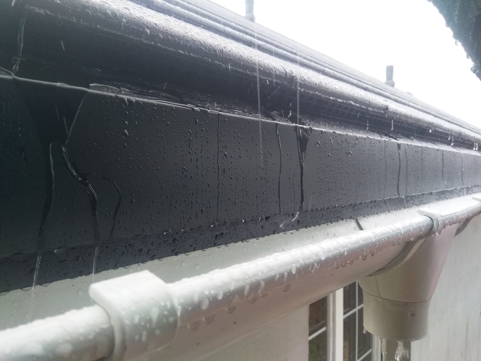 Czarna, mokra podsufitka dachu i biała rynna podczas deszczu, krople wody spływają po elementach systemu odprowadzania wody.