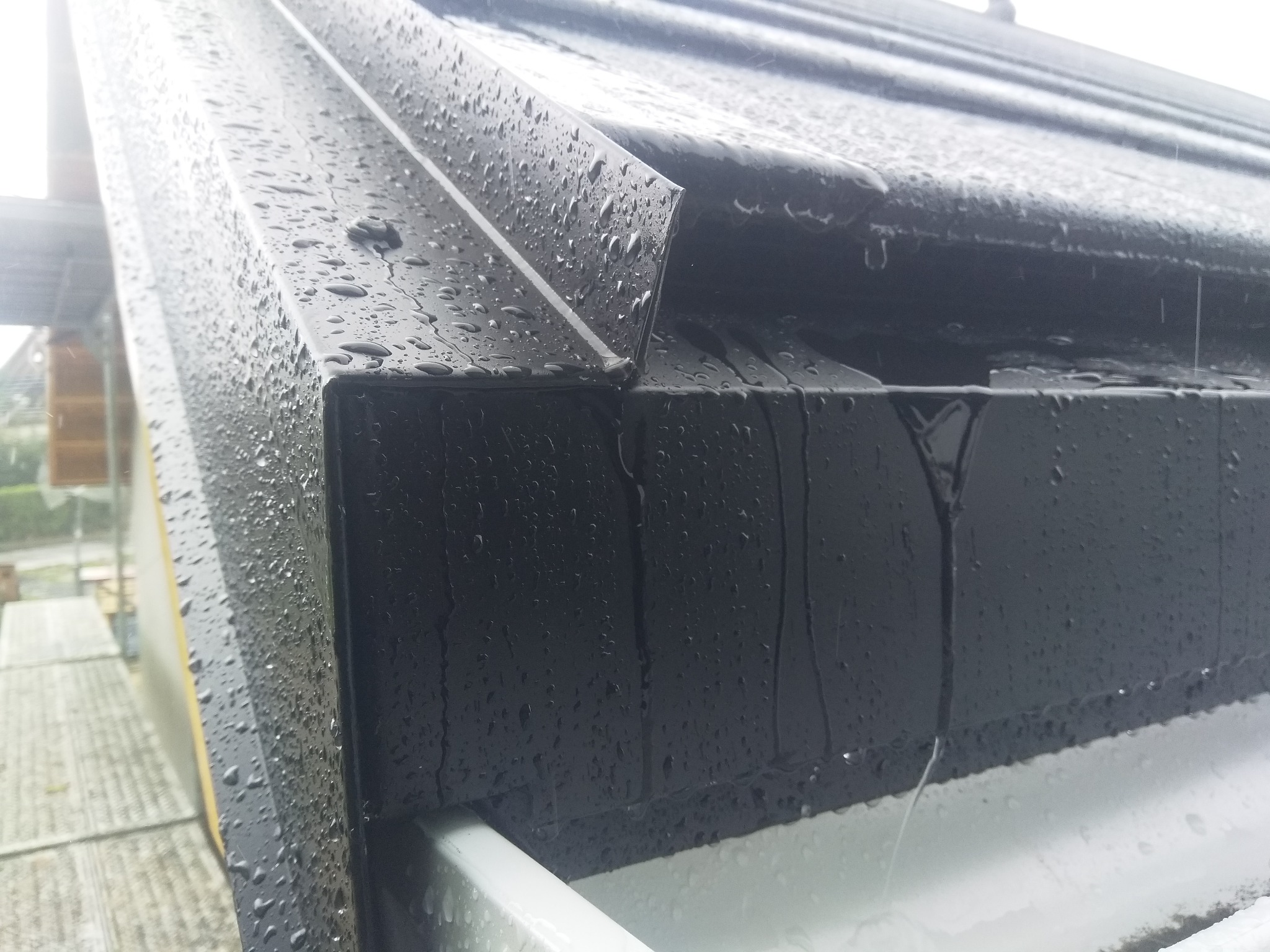 Czarny, metalowy fragment dachu z widoczną rynną, pokryty kroplami deszczu, sfotografowany z bliska pod kątem, z widocznym fragmentem rusztowania w tle.