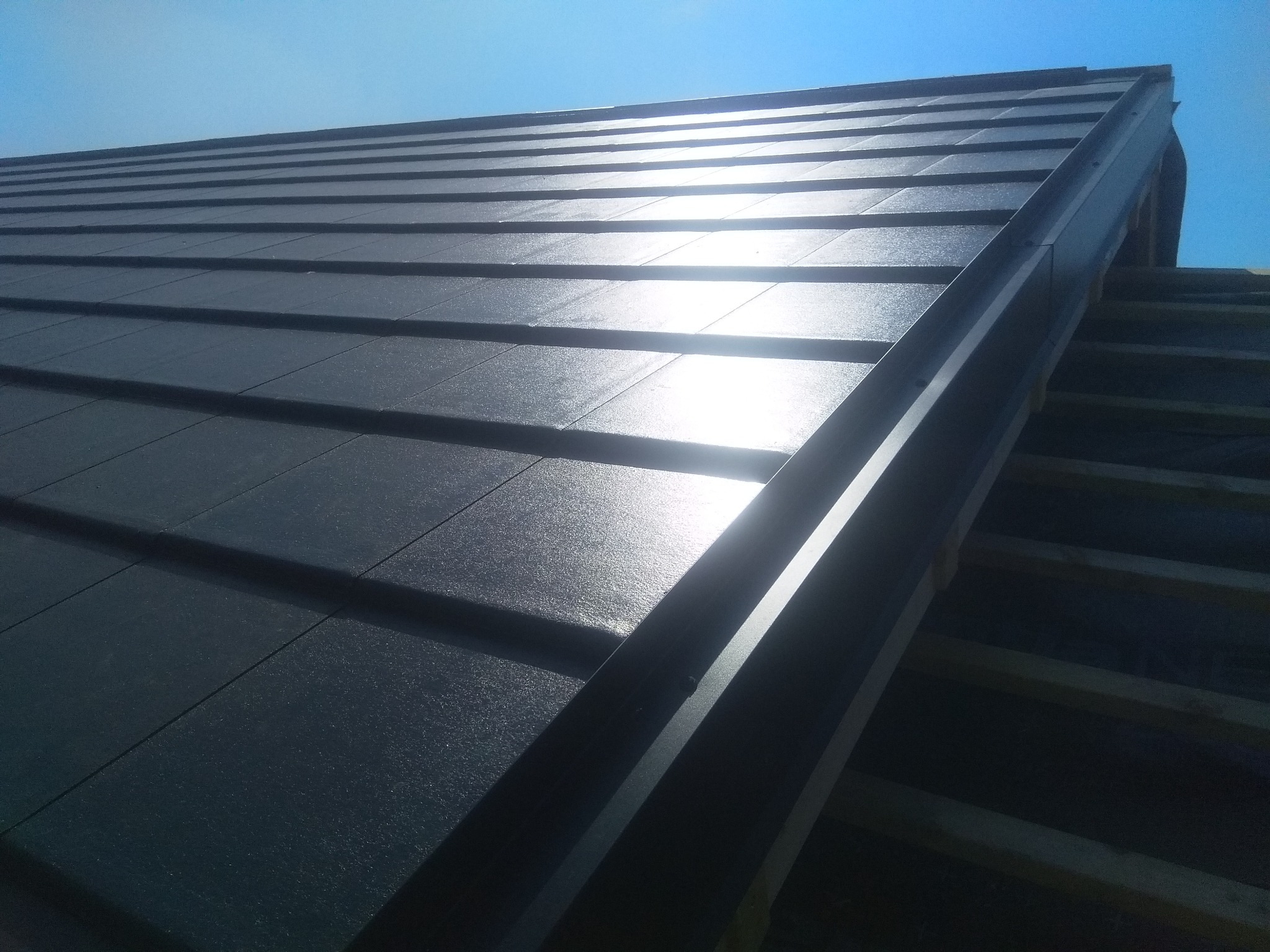 Nowy, ciemny dach z blachy na drewnianej konstrukcji, oświetlony jasnym słońcem, widok z dołu.