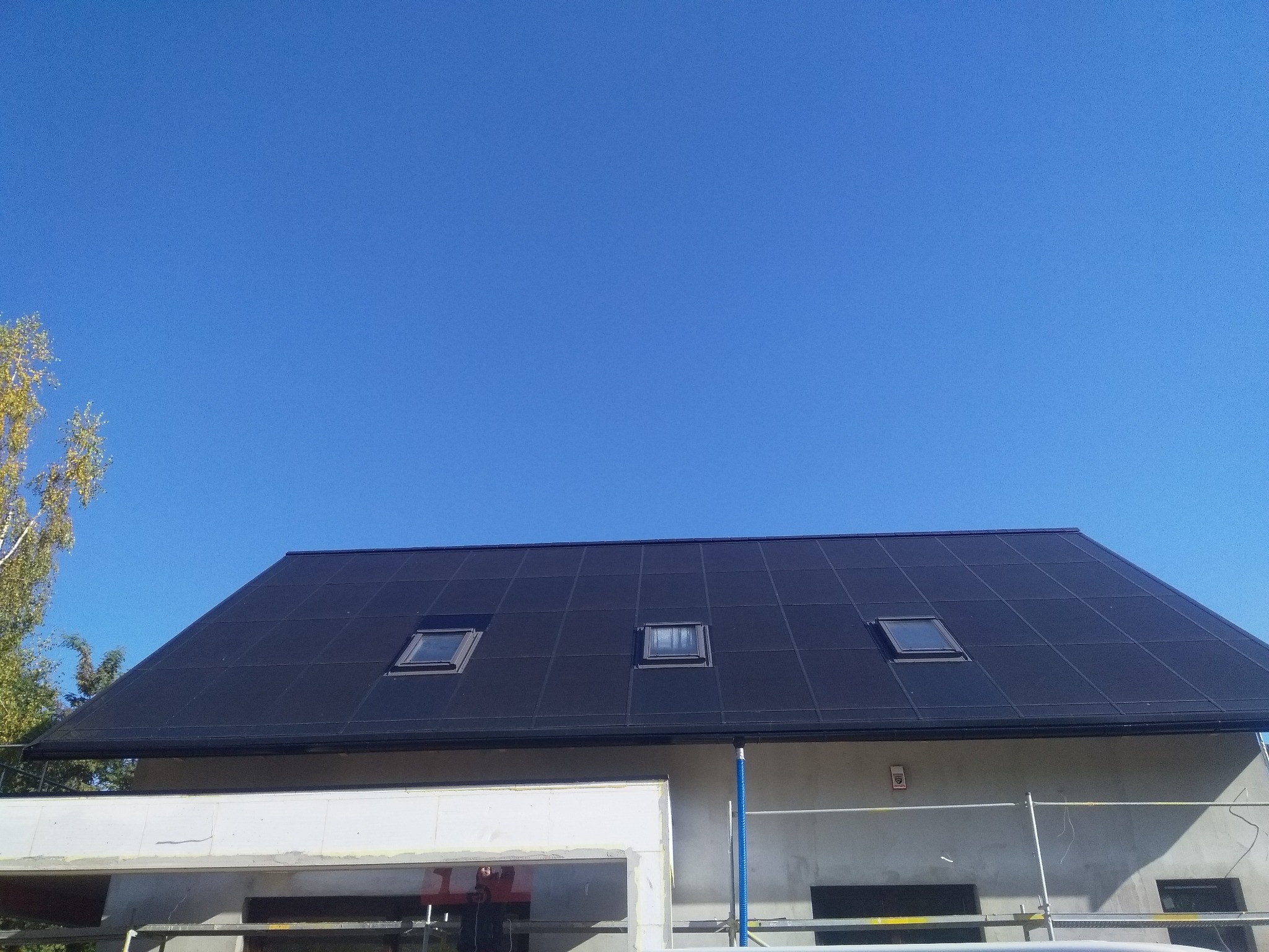 Czarny dach z trzema oknami dachowymi na tle błękitnego nieba, budynek w trakcie budowy z widocznymi rusztowaniami.