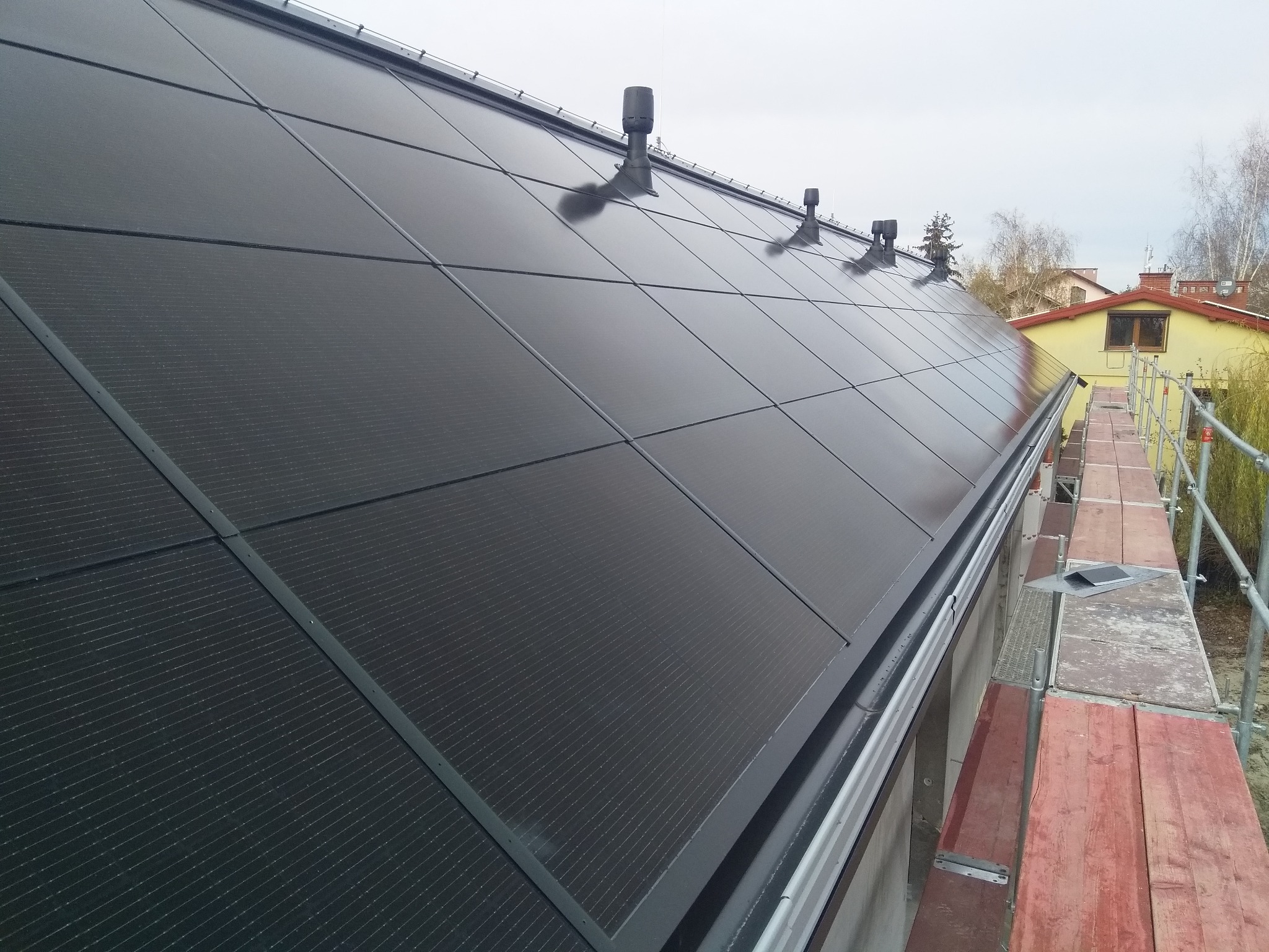 Czarny, matowy dach z panelami fotowoltaicznymi, widoczne kominki wentylacyjne i system rynnowy, ujęcie z rusztowania.