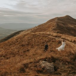 Photo Mood Paulina Bogon - Para młoda wędruje ścieżką na szczycie góry porośniętej suchą trawą, w tle widoczne zalesione wzgórza i pochmurne niebo.