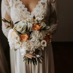 Panna młoda w koronkowej sukni trzyma bukiet ślubny z białych i pomarańczowych róż oraz goździków, widoczne dłonie z jasnym manicure.