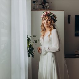 Photo Mood Paulina Bogon - Uśmiechnięta panna młoda w białej sukni z długimi rękawami i wiankiem z kwiatów na głowie, stojąca przy oknie z firanką, obok zielonej rośliny doniczkowej.