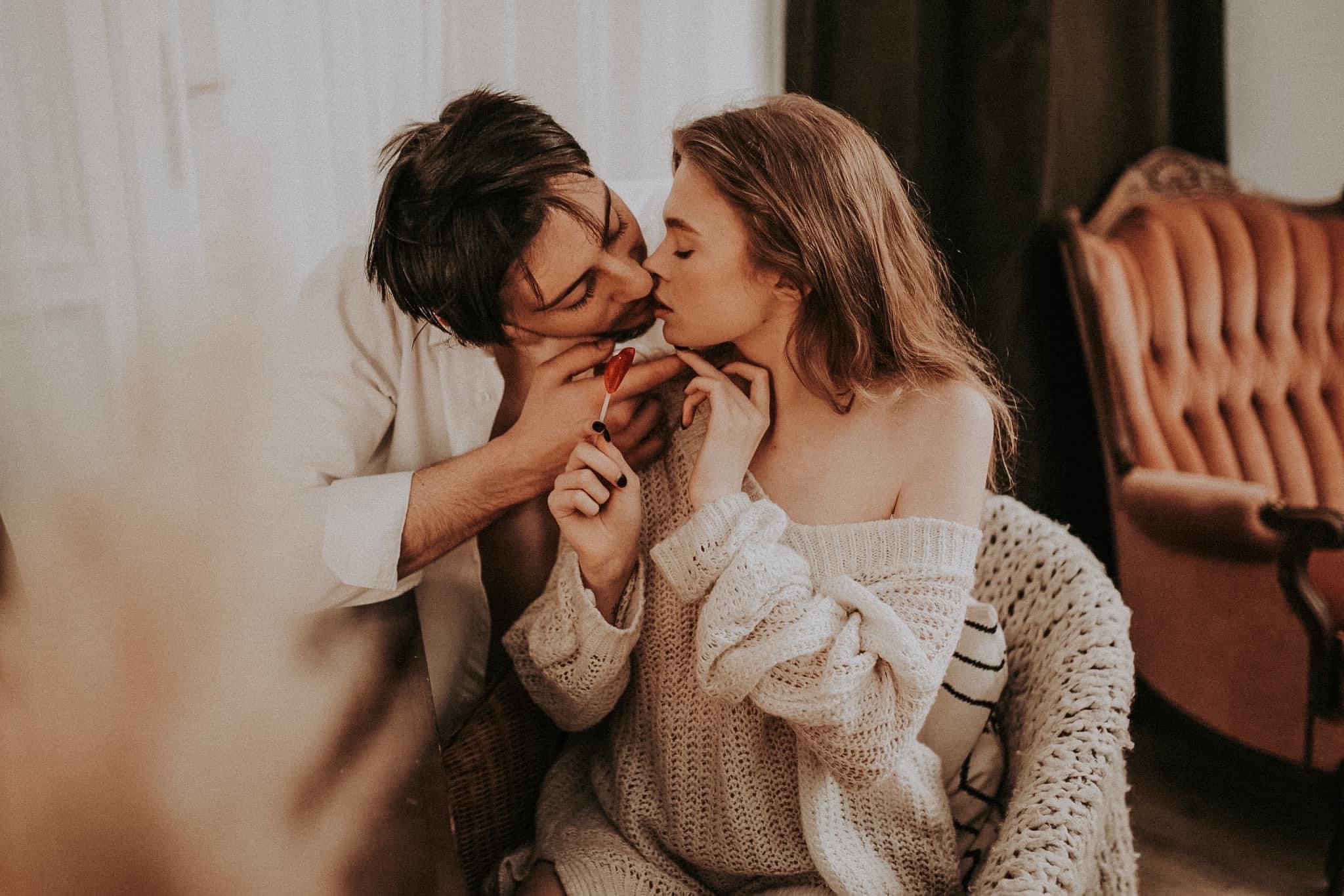 Para całująca się, trzymająca lizaka w kształcie serca, ona w grubym swetrze z odkrytymi ramionami, siedzi w wiklinowym fotelu, w tle fotel w stylu vintage