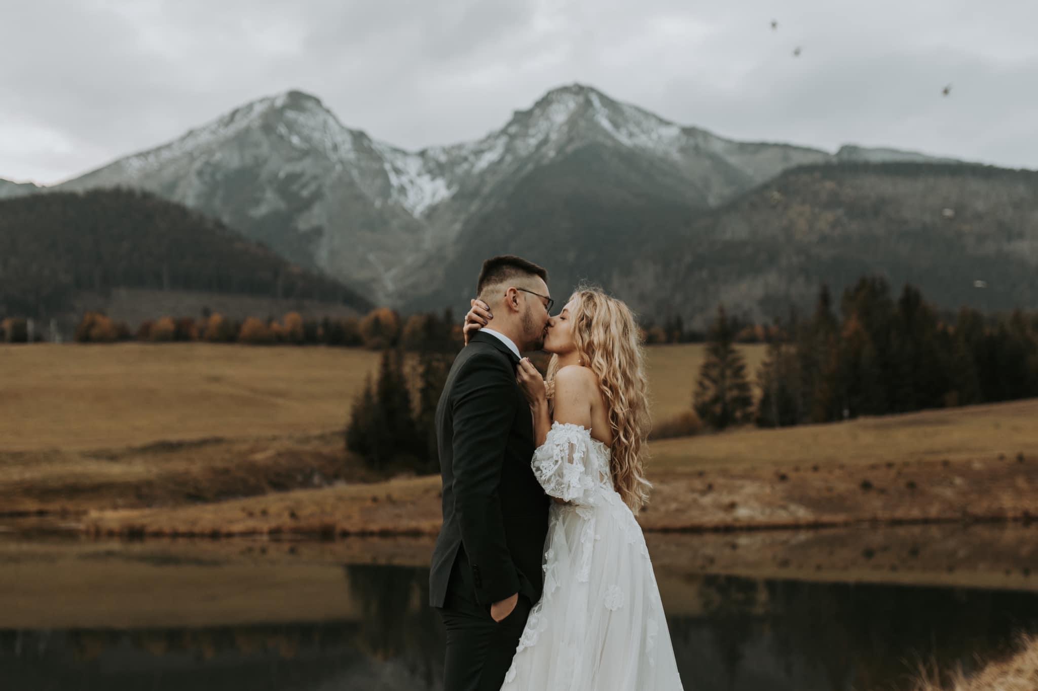 Pocałunek pary młodej na tle gór z ośnieżonymi szczytami, panna młoda w sukni z koronkowymi rękawami, pan młody w czarnym garniturze i okularach.