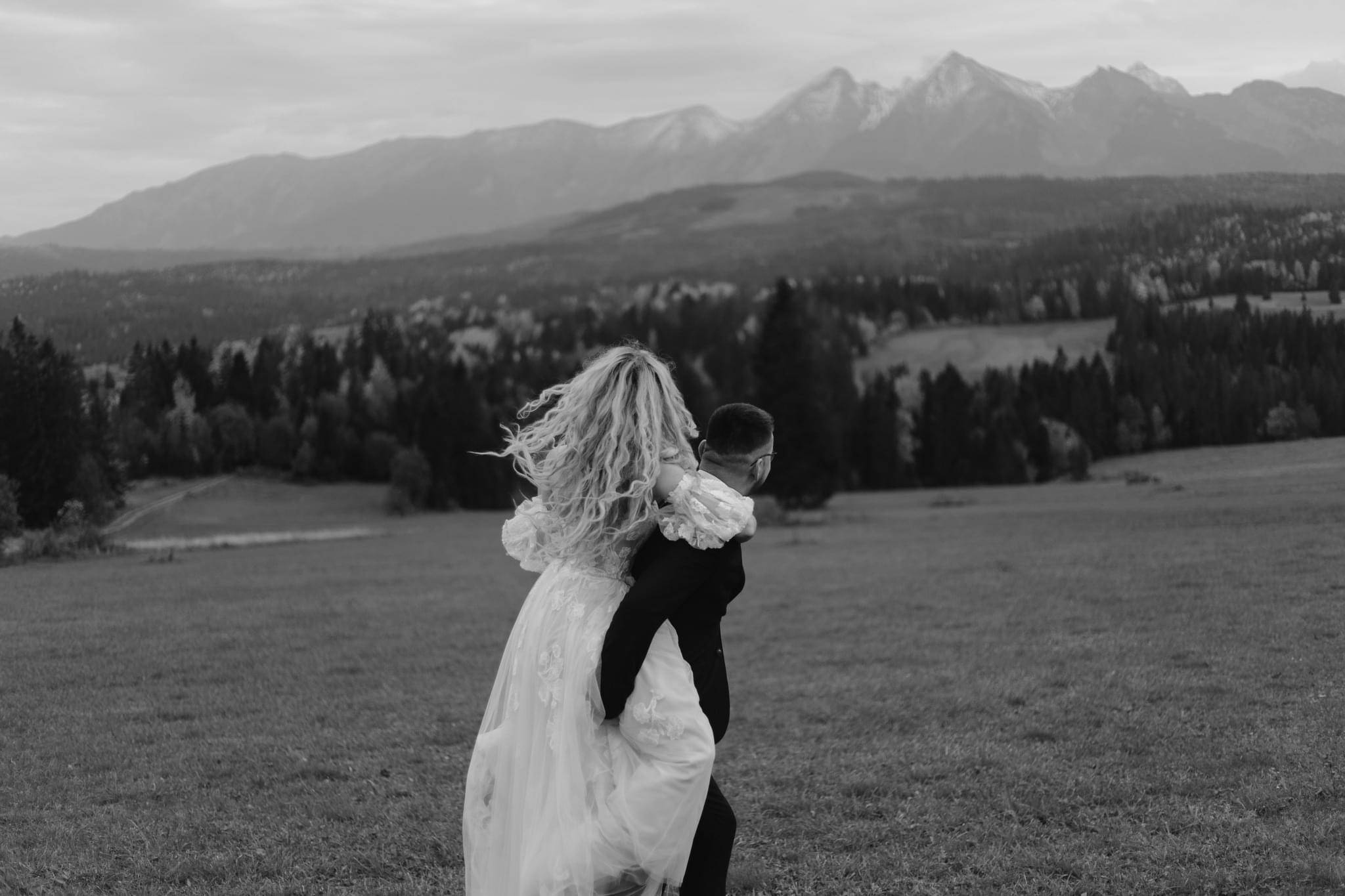 Pan młody niesie pannę młodą na plecach na tle gór, zdjęcie czarno-białe, panna młoda ma długie blond włosy, widok z tyłu.