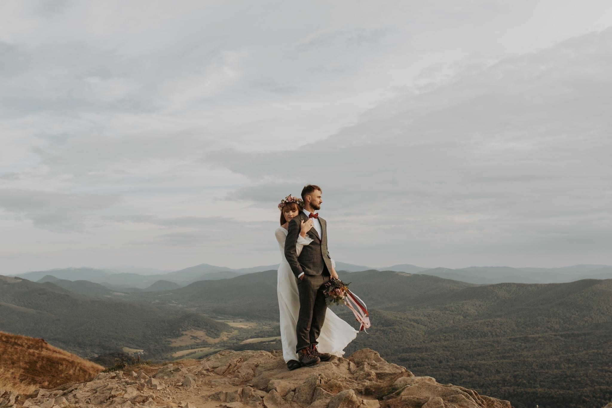 Para młoda w strojach ślubnych stoi na szczycie góry z bukietem, na tle rozległych górskich krajobrazów i pochmurnego nieba.