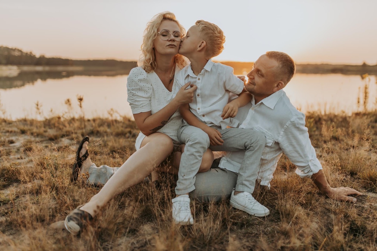 Rodzinne zdjęcie w Tarnowie: Mama w białej sukience i okularach siedzi na trawie z synem, który całuje ją w policzek, obok tata w białej koszuli, w tle jezioro i zachodzące słońce.