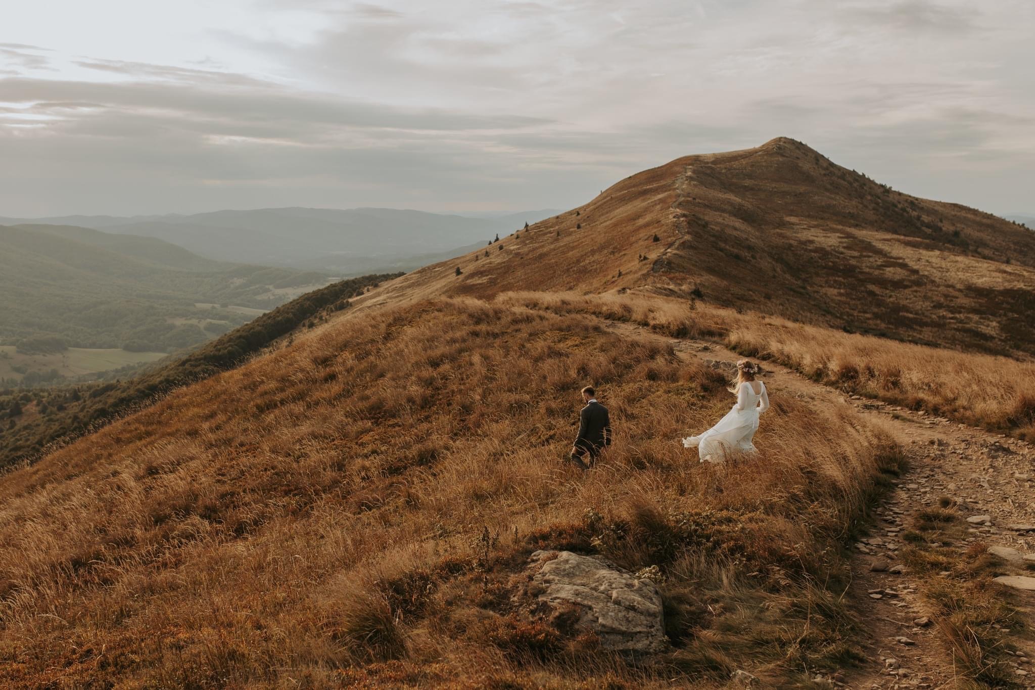 Para młoda wędruje ścieżką na szczycie góry porośniętej suchą trawą, w tle widoczne zalesione wzgórza i pochmurne niebo.