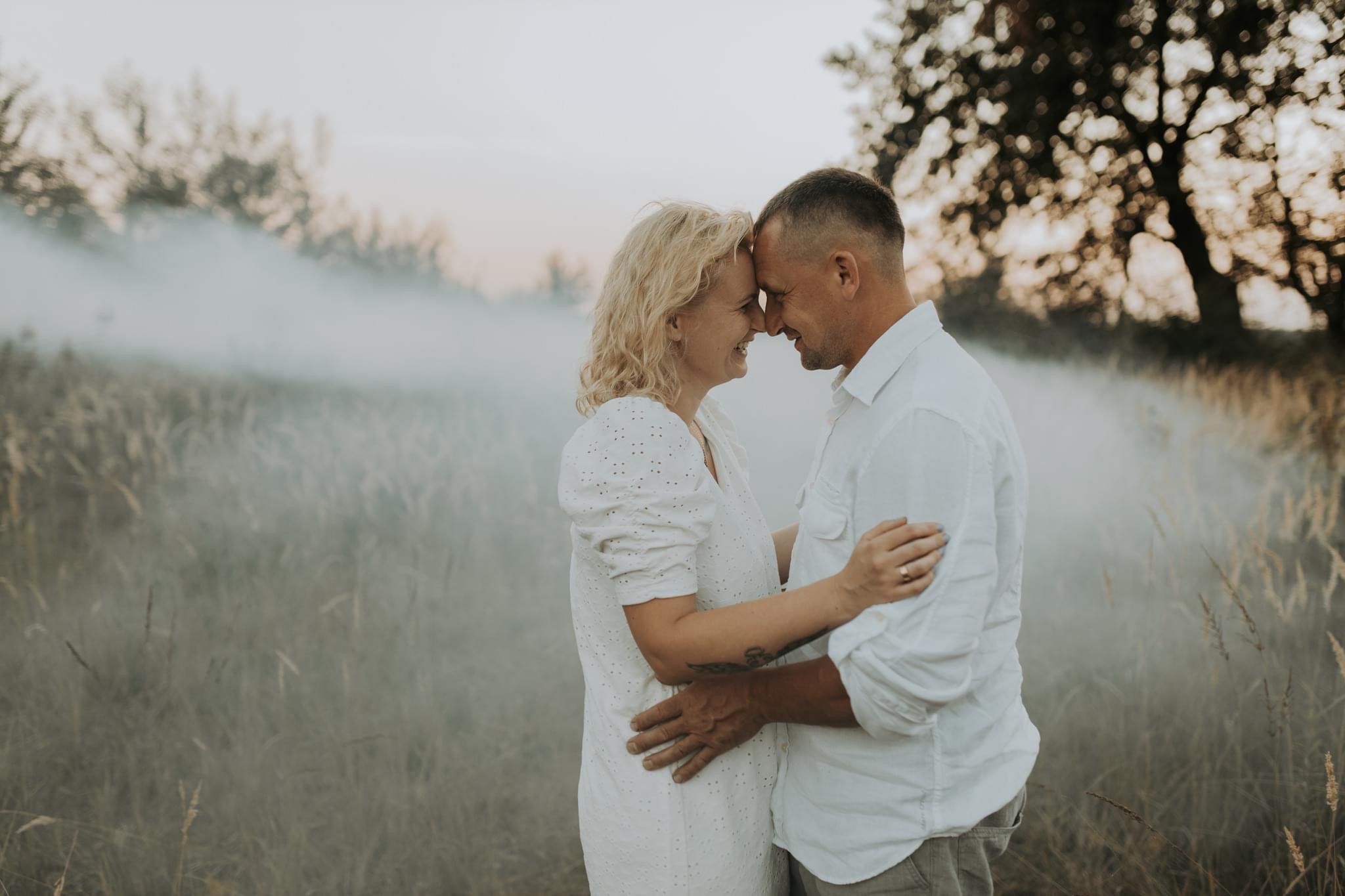 Uśmiechnięta para, kobieta w ażurowej białej sukience i mężczyzna w białej koszuli, przytuleni czołami na tle łąki z mgłą.