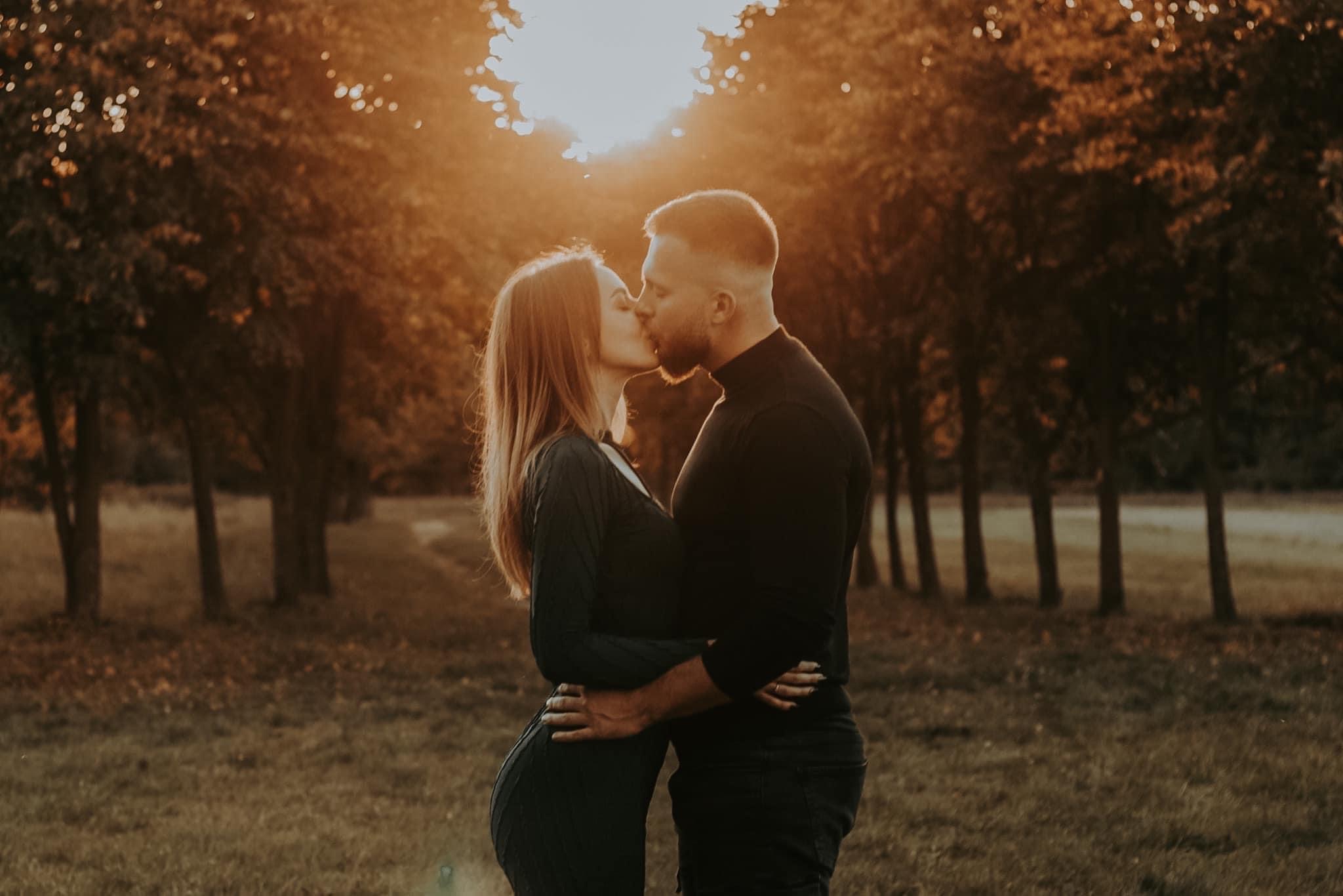 Zakochana para w czarnych ubraniach całuje się na tle drzew oświetlonych zachodzącym słońcem, tworzącym złocistą aureolę.