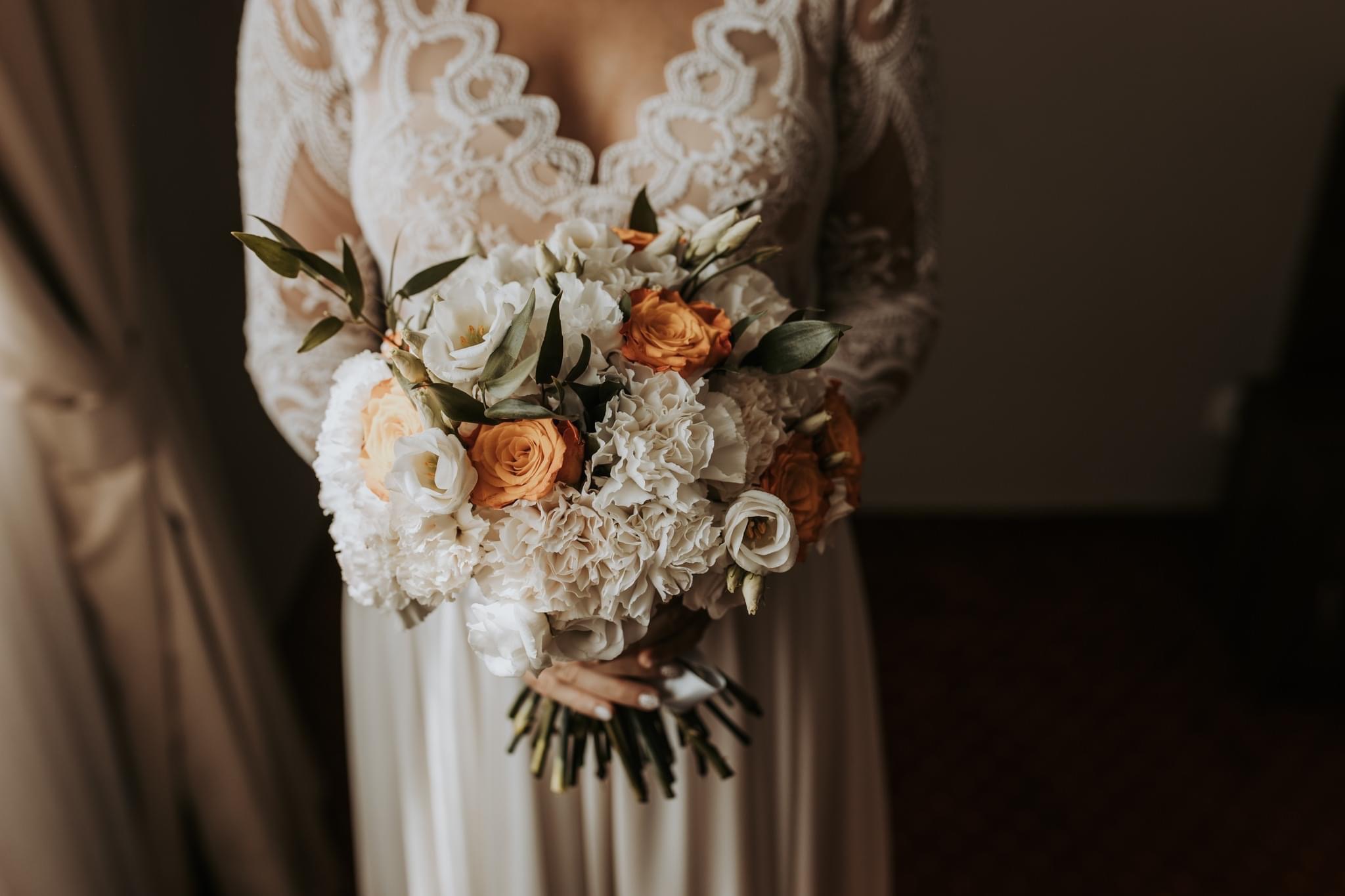 Panna młoda w koronkowej sukni trzyma bukiet ślubny z białych i pomarańczowych róż oraz goździków, widoczne dłonie z jasnym manicure.