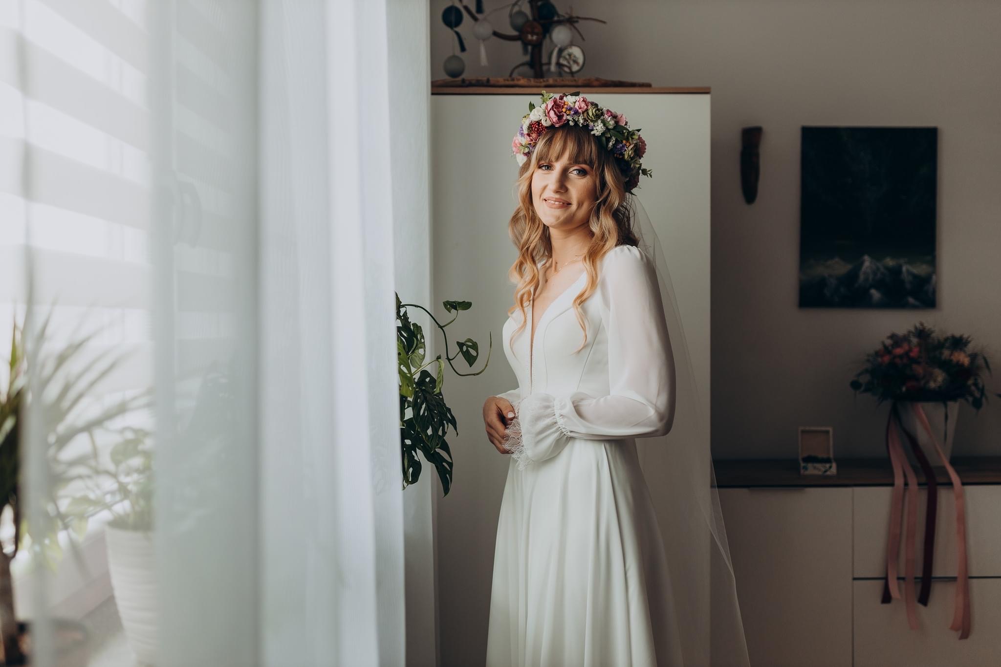 Uśmiechnięta panna młoda w białej sukni z długimi rękawami i wiankiem z kwiatów na głowie, stojąca przy oknie z firanką, obok zielonej rośliny doniczkowej.