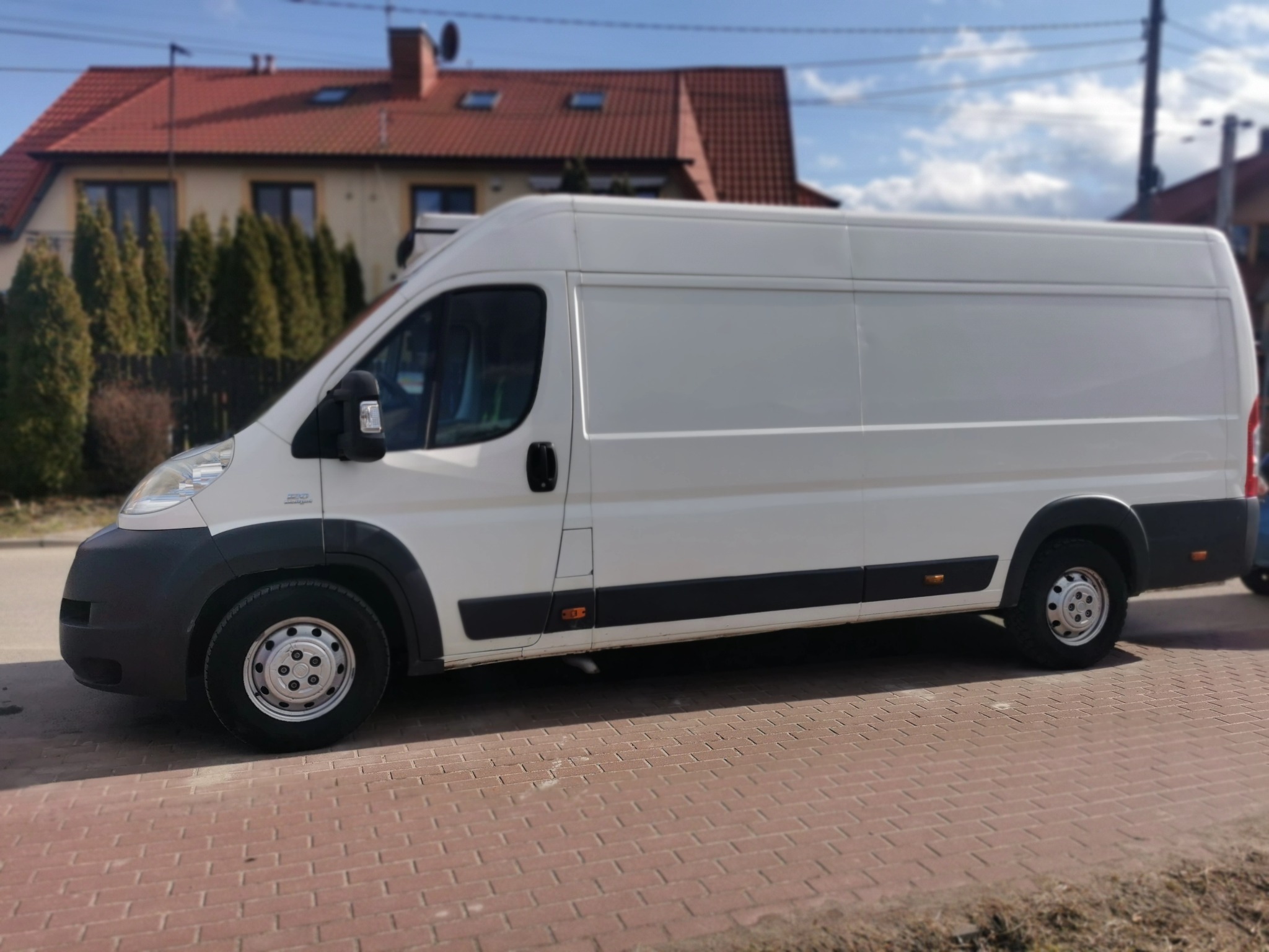 Biały samochód dostawczy Fiat Ducato z wysokim dachem zaparkowany na czerwonej kostce brukowej przed domem z czerwoną dachówką.