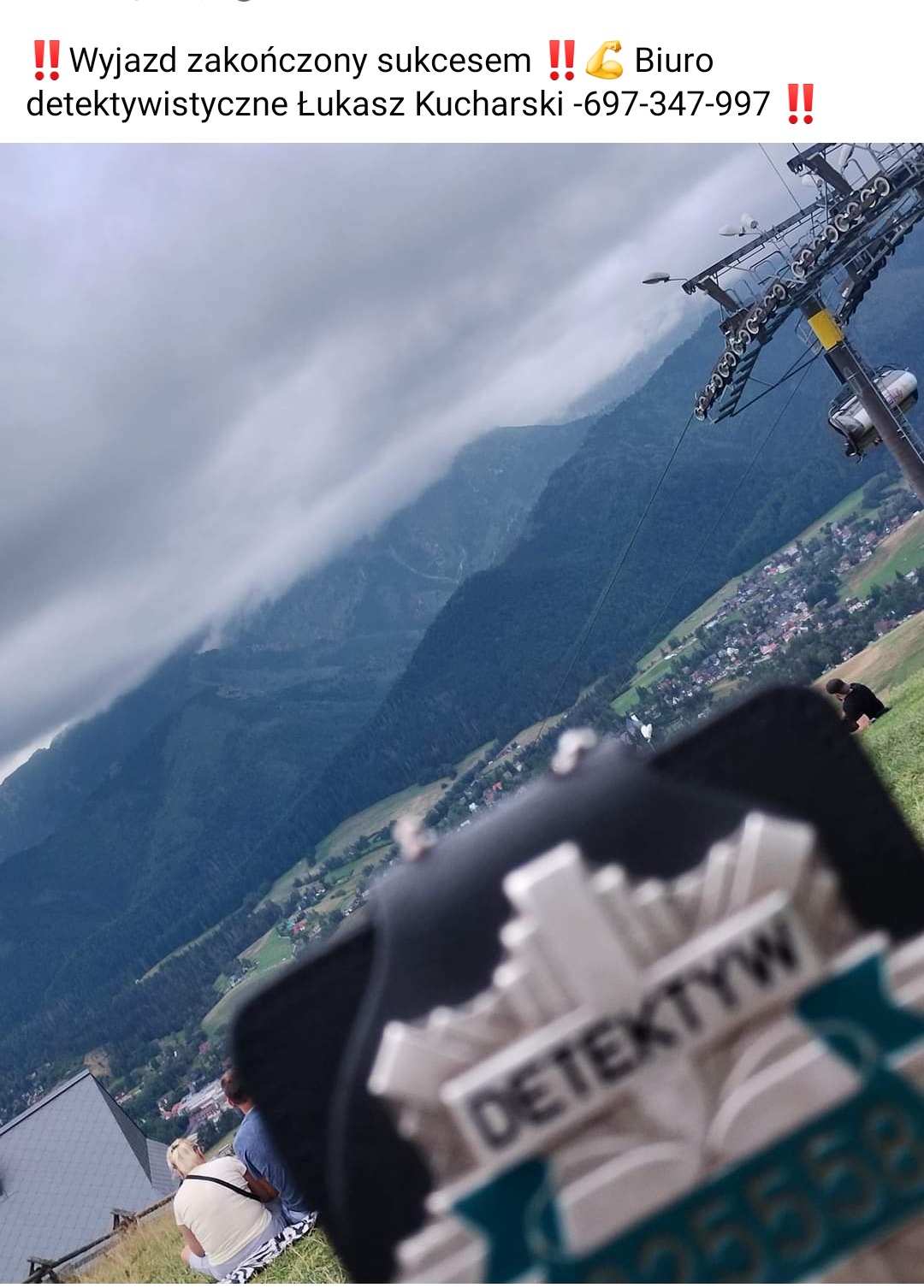 Górski krajobraz z widoczną kolejką linową, u dołu kadru naklejka z napisem 'Detektyw' i numerem seryjnym, osoby siedzące na trawie obserwujące otoczenie.