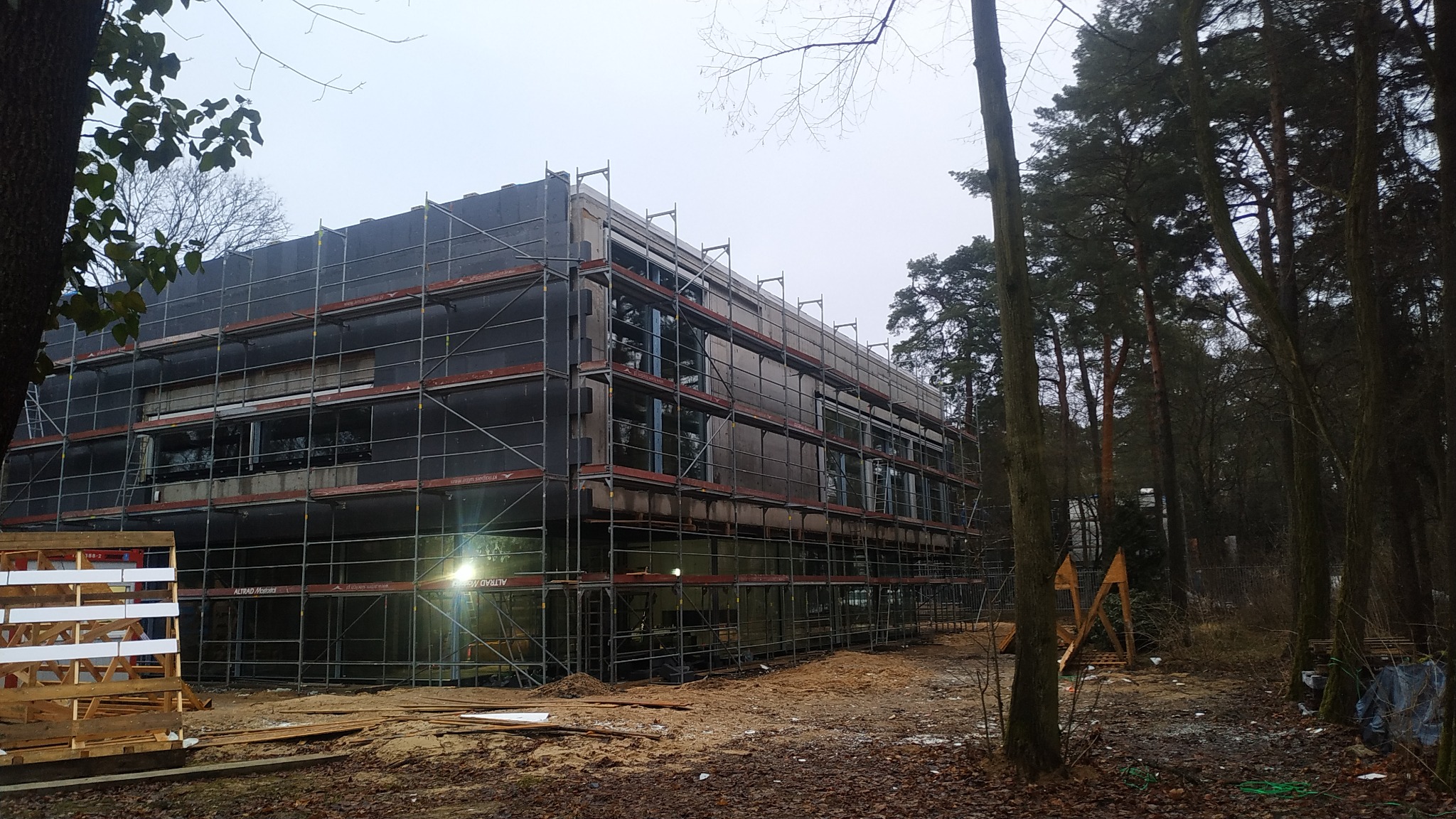 Budynek w trakcie prac budowlanych, z rusztowaniami i częściowo ocieploną elewacją, otoczony drzewami i ziemią z liśćmi.
