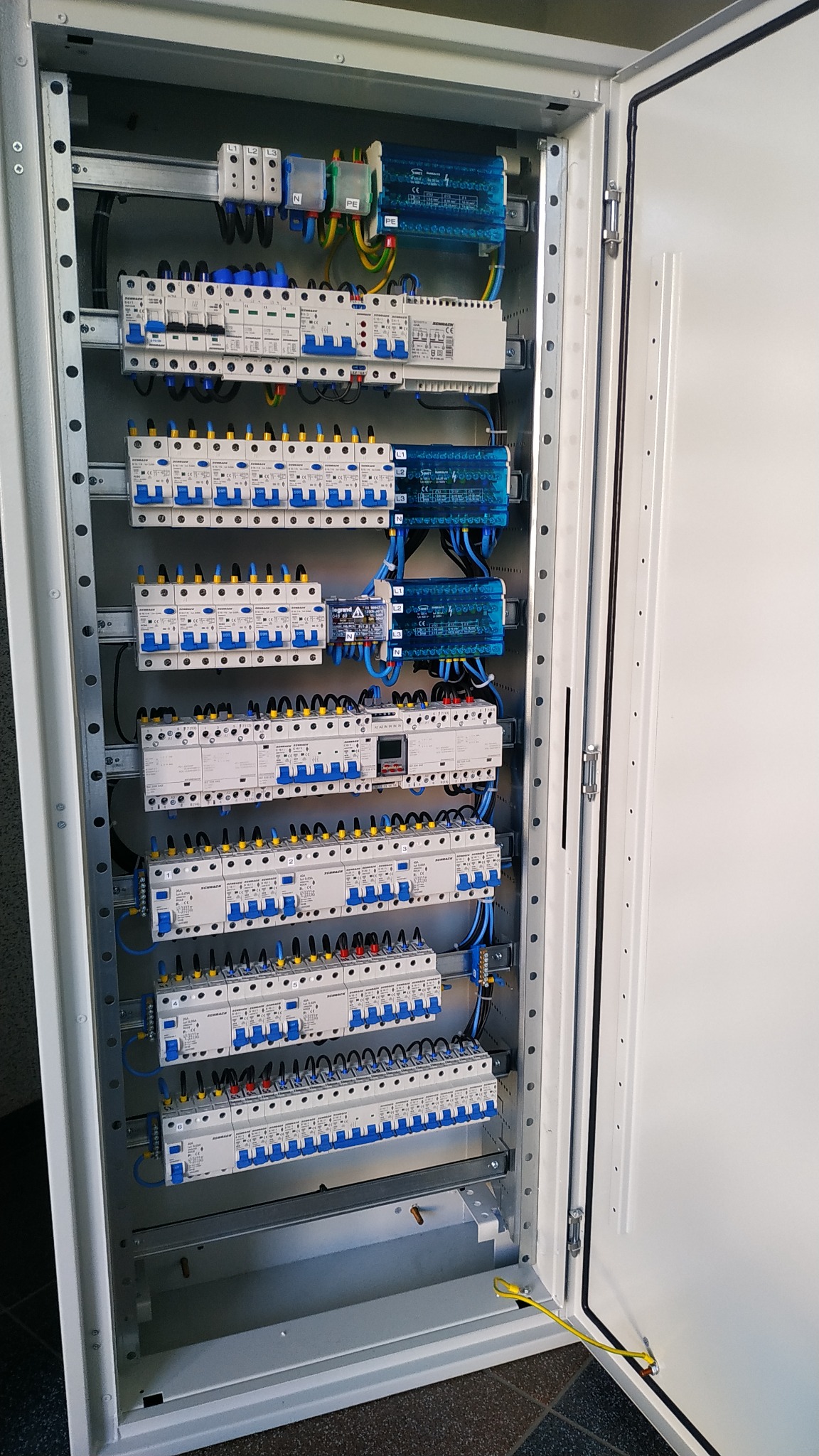 Otwarta skrzynka elektryczna z zamontowanymi bezpiecznikami, modułami i okablowaniem. Widoczne przewody w kolorach żółto-zielonym, niebieskim i czarnym. Metalowa obudowa z szyną DIN.