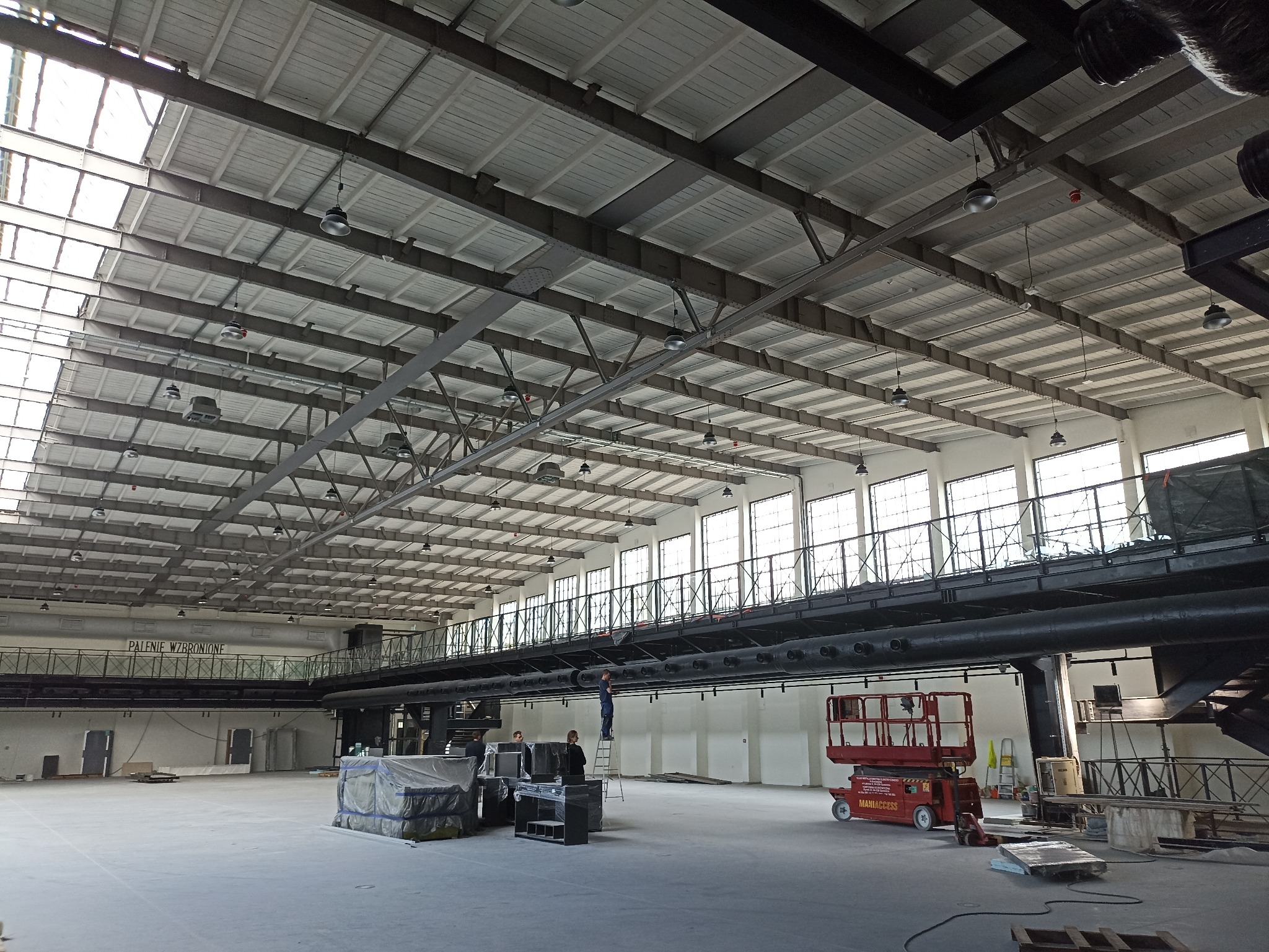Wnętrze hali z widoczną konstrukcją dachu, oświetleniem przemysłowym i pracownikami na podnośniku oraz drabinie, wykonującymi prace na wysokości.