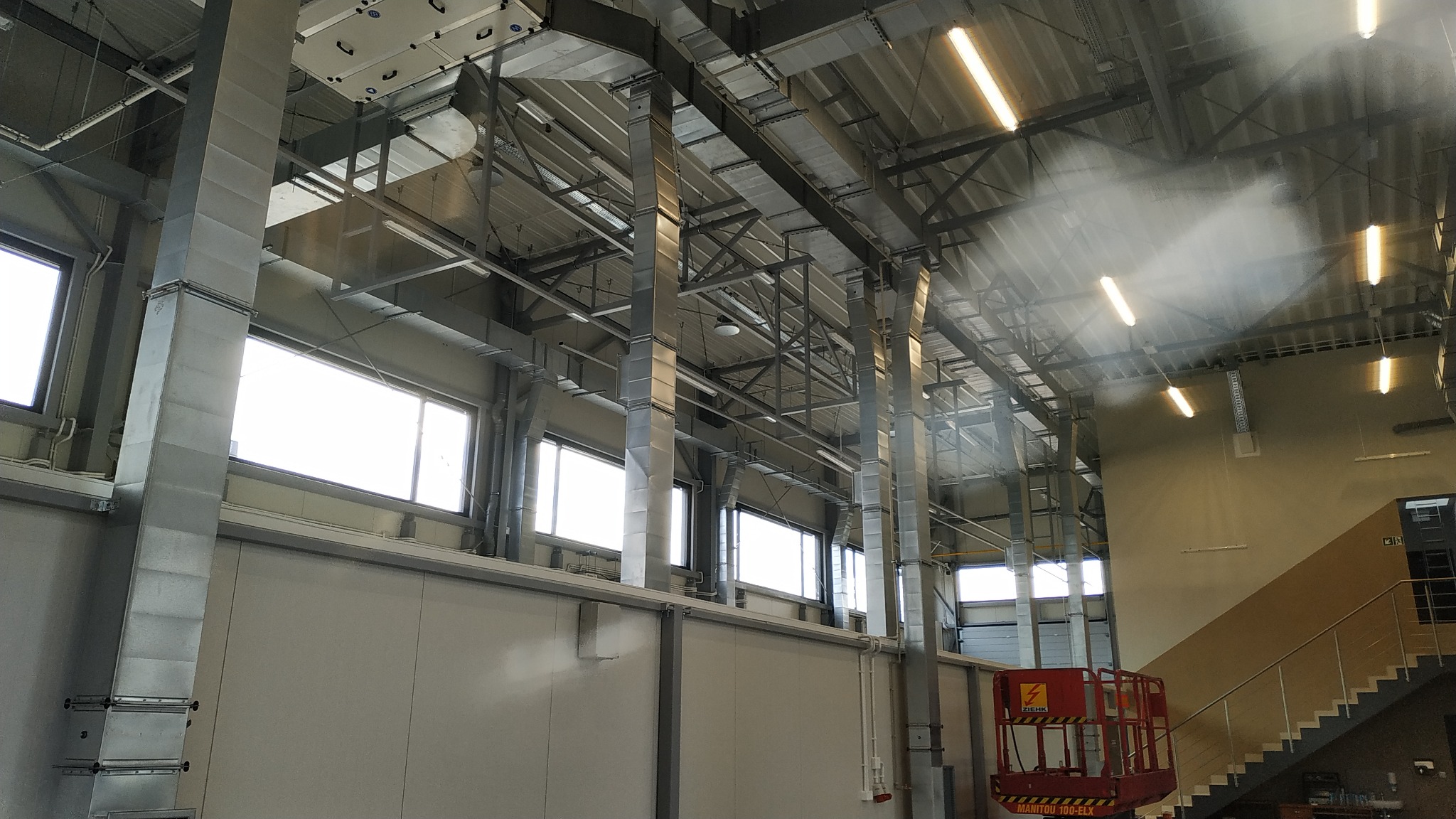 Wnętrze hali przemysłowej z widocznymi elementami wentylacji, klimatyzacji i instalacji elektrycznych, w tym kanały wentylacyjne, kolumny, okna i oświetlenie sufitowe. W tle widoczna winda towarowa.