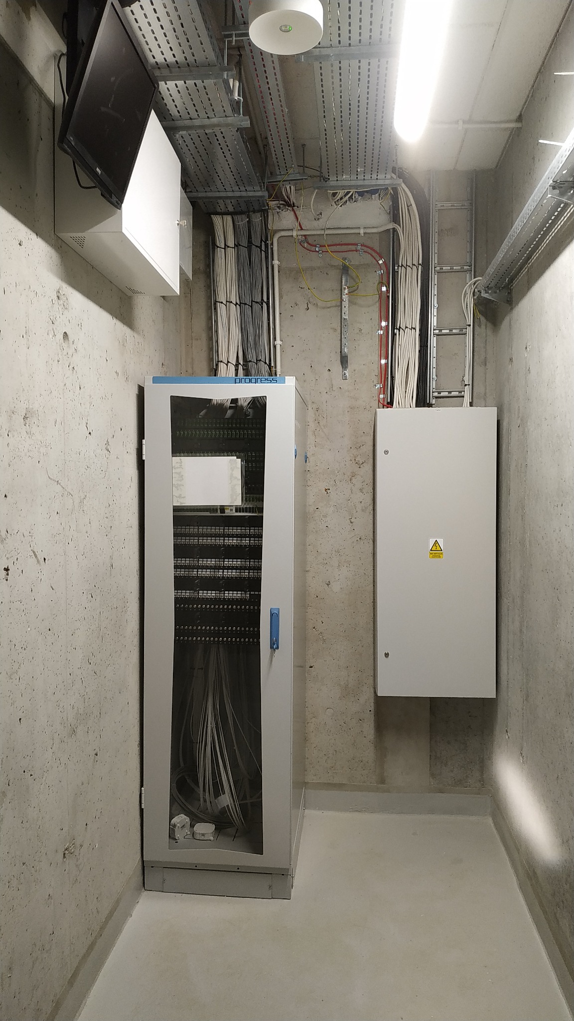 Wnętrze pomieszczenia technicznego z szafą telekomunikacyjną, rozdzielnicą elektryczną, korytami kablowymi pod sufitem i przewodami ułożonymi w wiązki.