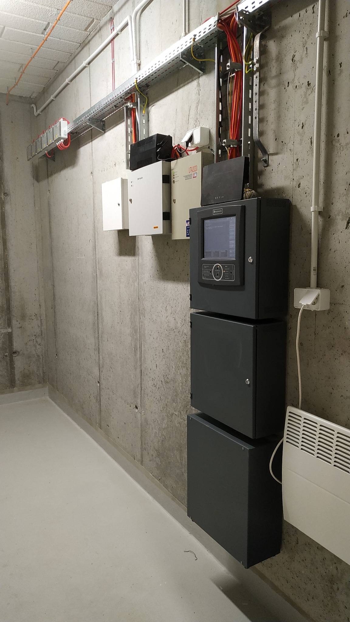 Szara szafa sterownicza z ekranem dotykowym i trzema modułami, zamontowana na surowej betonowej ścianie w pomieszczeniu technicznym, widoczne kable w korytkach i biały grzejnik konwektorowy.