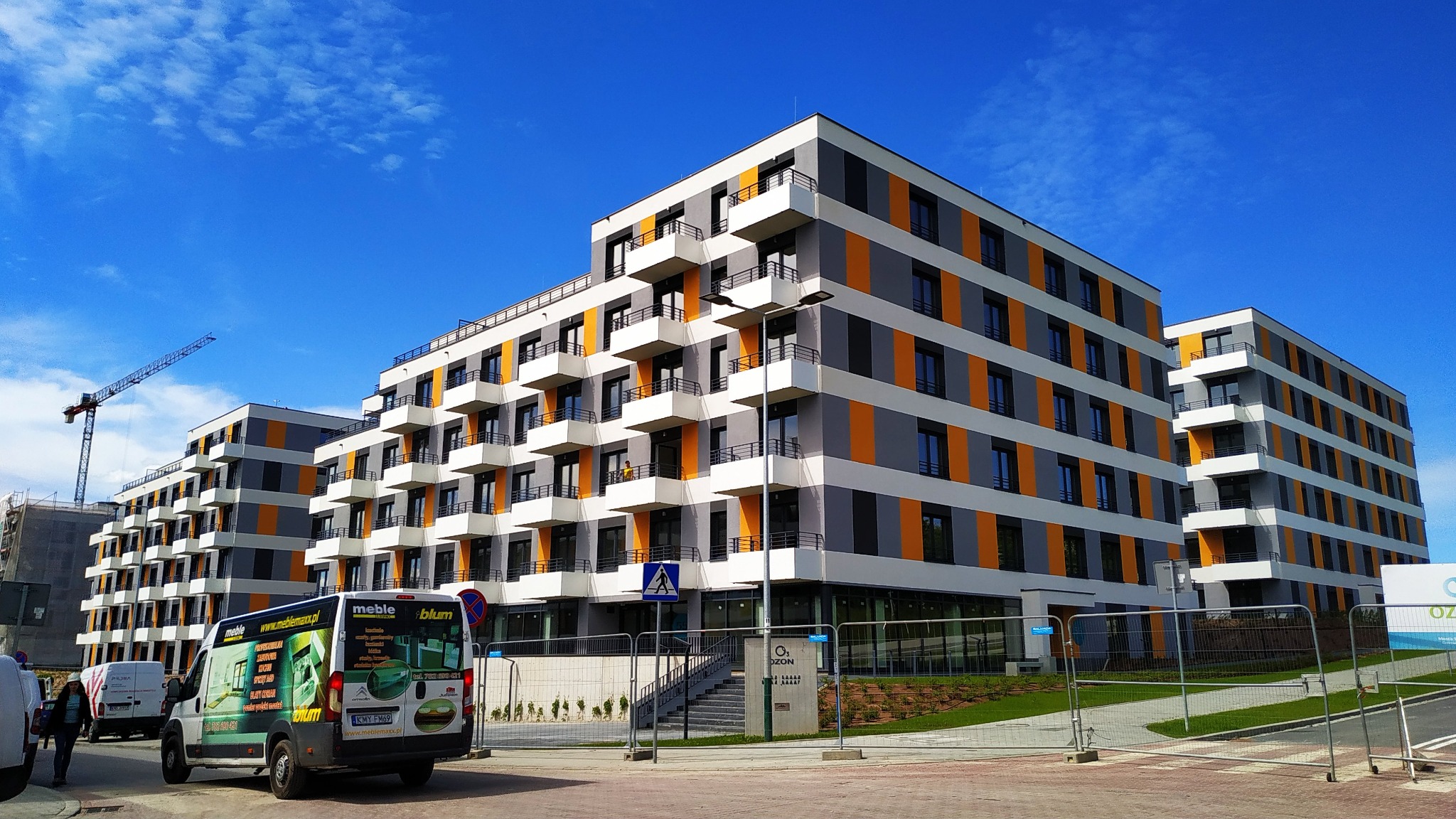 Nowoczesne budynki mieszkalne z balkonami w Krakowie, w tle żuraw budowlany, na pierwszym planie samochód dostawczy firmy meblowej z reklamą blatów kuchennych.