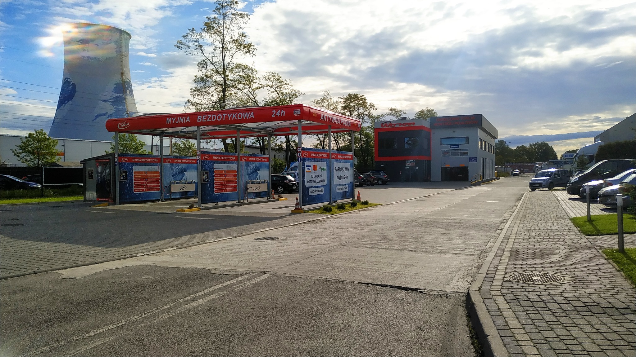 Widok na samoobsługową myjnię bezdotykową z czerwonym zadaszeniem i budynkiem obsługi w tle, w oddali widoczna chłodnia kominowa.