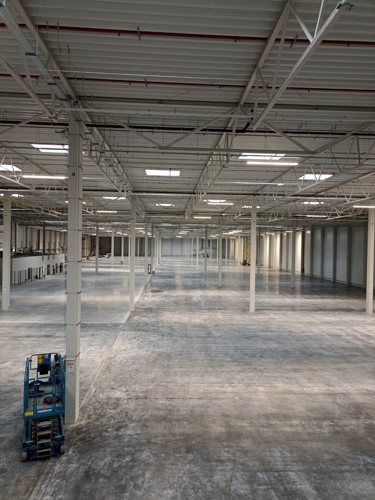 Wnętrze pustej hali magazynowej z widoczną konstrukcją dachu, oświetleniem, betonową posadzką i podnośnikiem koszowym w lewym dolnym rogu.