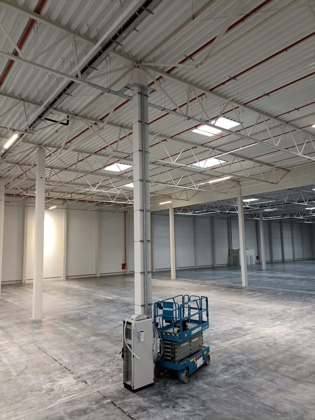 Wnętrze hali magazynowej z zamontowanymi instalacjami elektrycznymi pod sufitem, widoczny podnośnik nożycowy i szafa sterownicza.