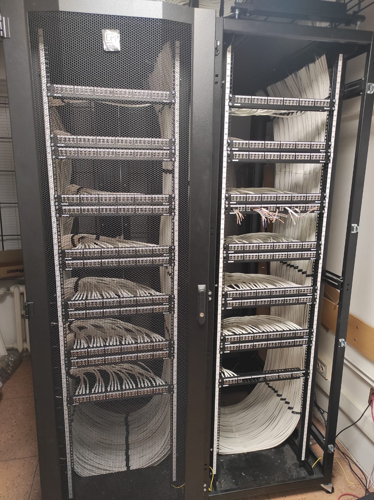 Dwie szafy rack z panelami krosowniczymi i uporządkowanymi kablami sieciowymi, widoczne metalowe ramy i siatki zabezpieczające, kable ułożone w estetyczne pętle.