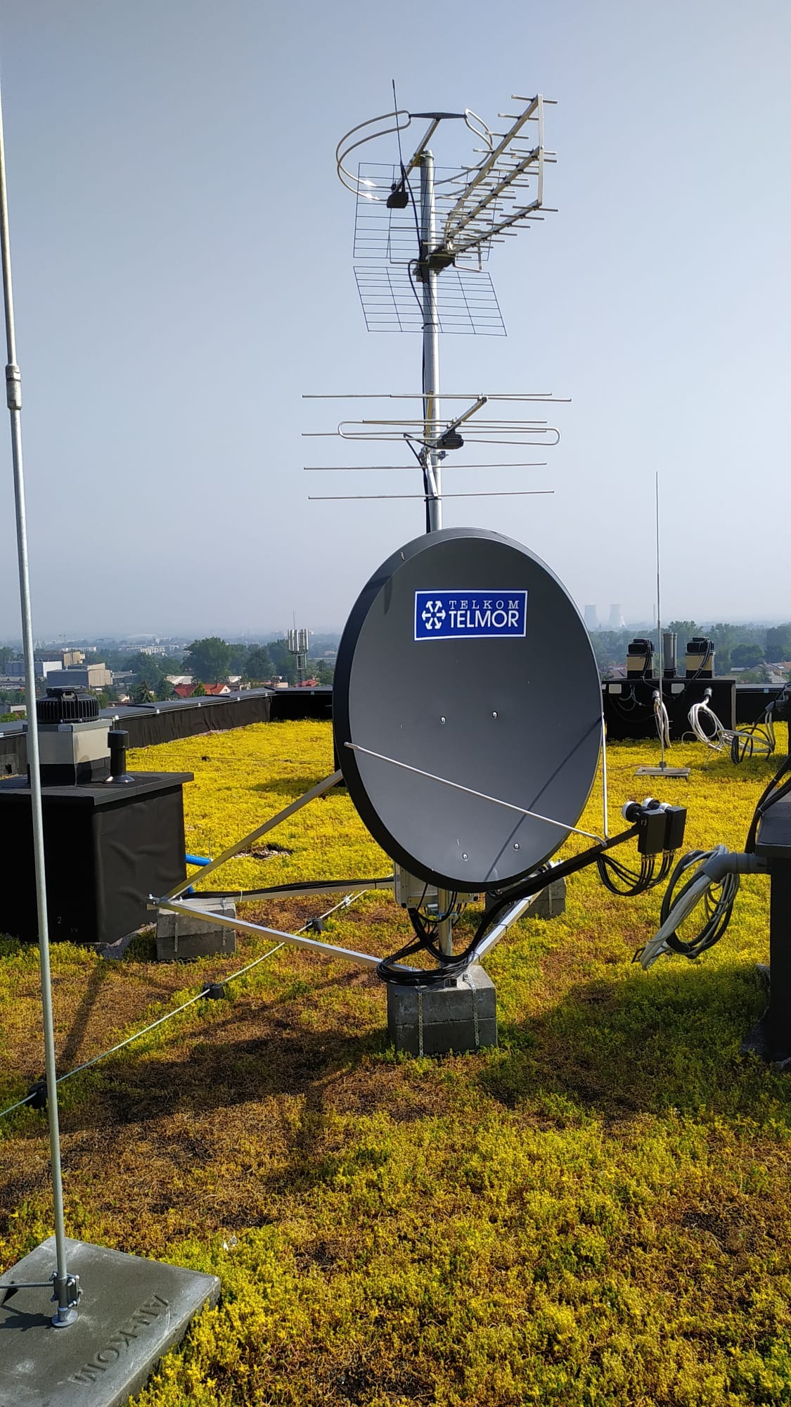 Antena satelitarna Telkom Telmor oraz antena kierunkowa na dachu pokrytym zielenią, w tle zabudowa miejska w lekkiej mgle.