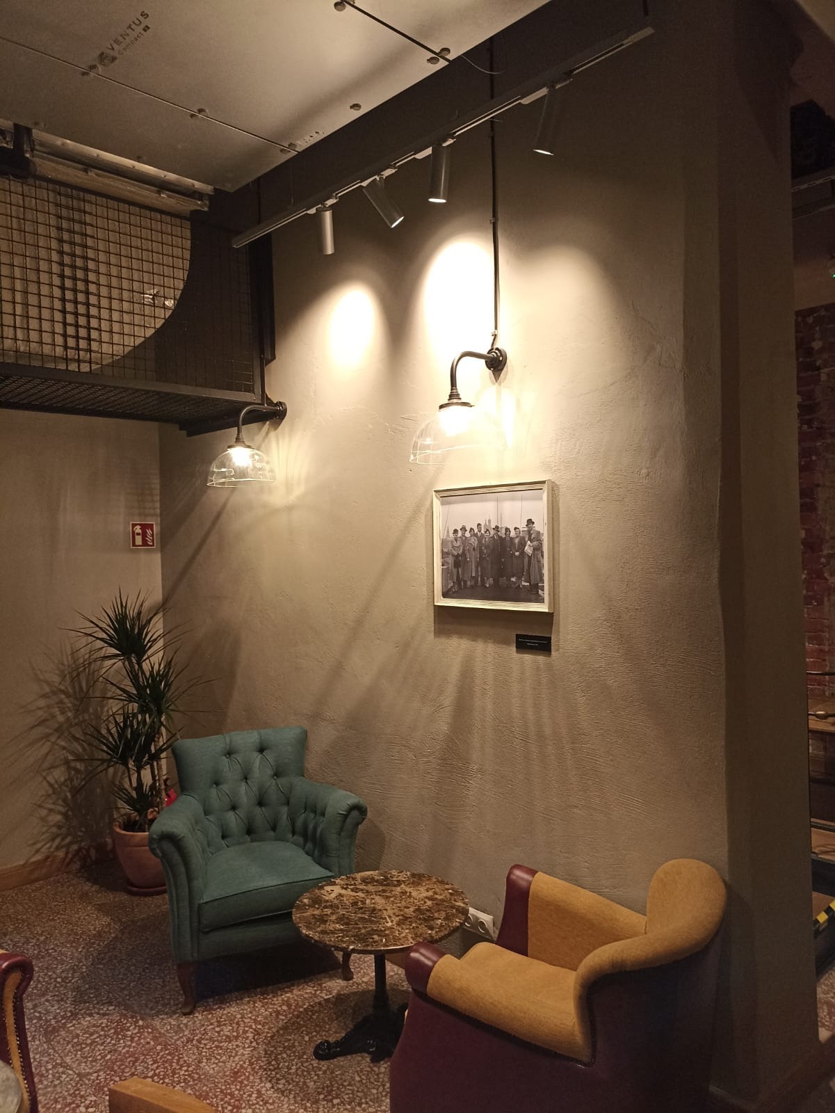 Stylowe wnętrze z oświetleniem punktowym na szynie, dwoma fotelami w różnych kolorach i małym okrągłym stolikiem. Na ścianie wisi czarno-białe zdjęcie pod industrialną antresolą z metalową siatką.